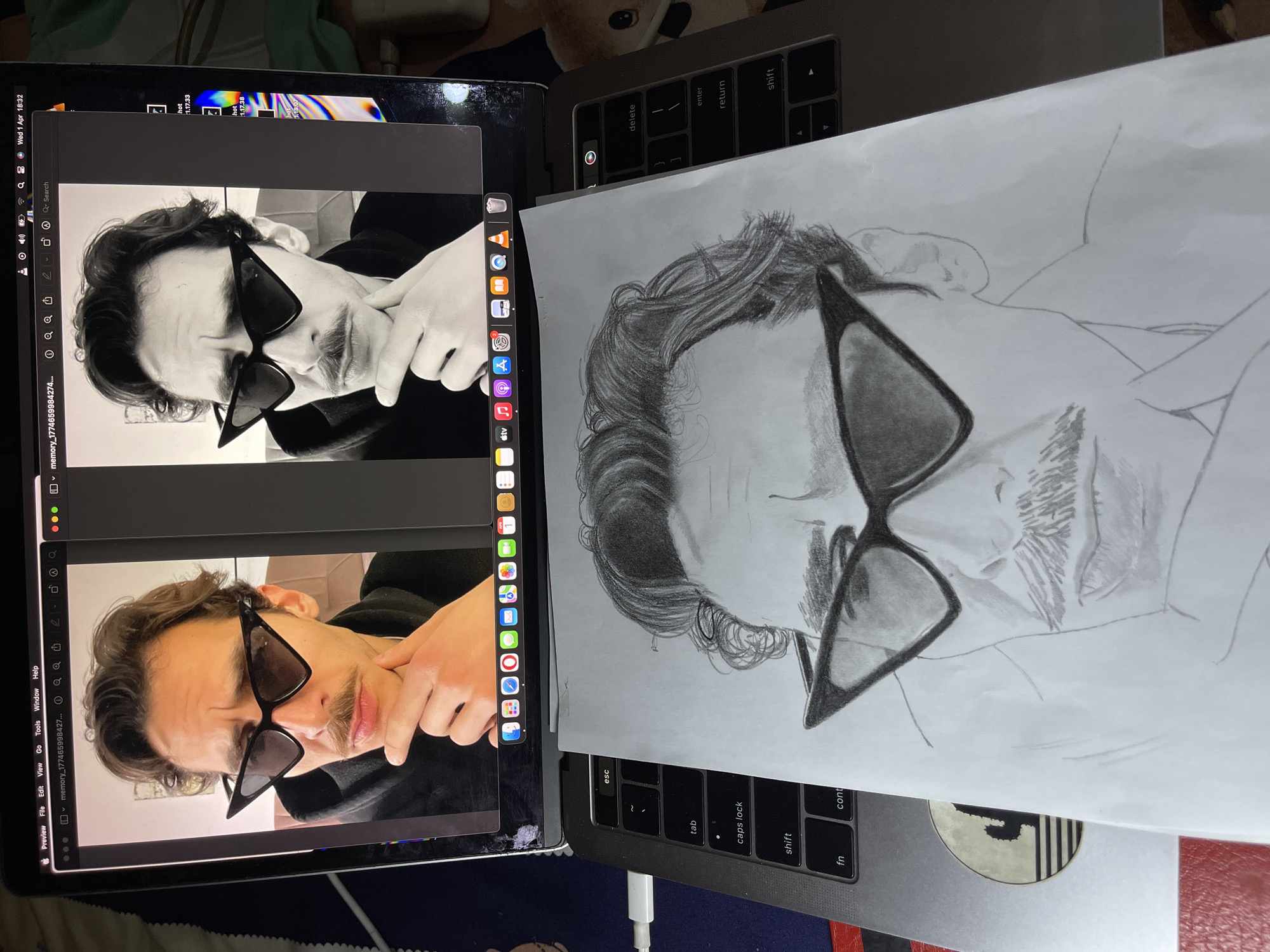 The image shows a partially completed charcoal or pencil drawing of a person wearing sunglasses, alongside a laptop. The laptop displays two similar images of the same person, also wearing sunglasses. The drawing rests on top of another laptop keyboard. The setting appears to be indoors, likely a home or studio given the casual arrangement and lack of identifiable features pinpointing a specific location in Jos, Nigeria, or any city. There are no people interacting in the scene, and the focus is on the drawing and its reference images on the laptop. The objects include the drawing, two laptops, and an unidentified sticker on the bottom right corner of the bottom laptop.