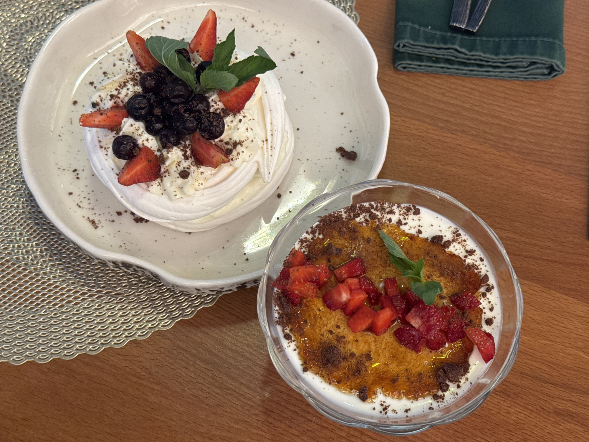 Two desserts are presented on a wooden table. In the foreground, a clear glass bowl contains a creamy dessert topped with caramelized sugar, chopped strawberries, and mint leaves. The caramelized sugar has a golden-brown hue, and scattered chocolate crumbs are visible on the cream. In the background, a Pavlova meringue dessert sits on a white plate. This dessert features a white meringue base, whipped cream, blueberries, sliced strawberries, and mint leaves, with chocolate crumbs sprinkled on top. A textured placemat with an intricate pattern is placed between the desserts and the table's edge. A dark green napkin, folded neatly, rests to the right of the desserts. The scene is captured from a top-down perspective, highlighting the desserts against the warm tones of the wooden surface. The environment is indoors, suggesting a dining setting in Sosnoviy Bor, Russia.