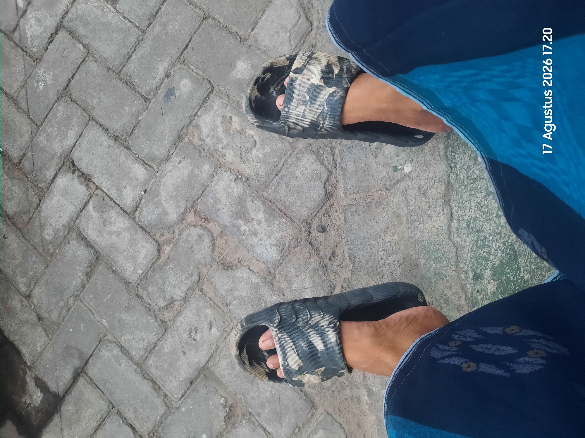 The image displays a pair of feet wearing camouflage-patterned slide sandals, standing on a paved walkway made of interlocking grey bricks. The feet are positioned close to the bottom of the frame, with the legs extending upwards and covered by dark blue fabric with a geometric pattern in lighter blue and white. The walkway has a textured surface with visible grout lines between the bricks. The lighting suggests an outdoor setting, likely during the day. The text overlay in the bottom right corner reads "17 Agustus 2026 17.20", indicating the date and time the image was captured, which is Wednesday, August 17, 2026, at 5:20 PM. The location of Sukokerto, Indonesia is not visually evident from the image itself.