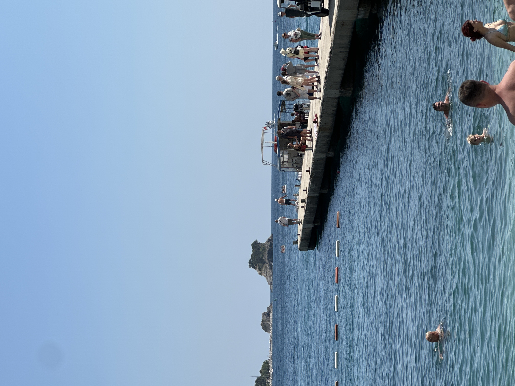 The image captures a sunny day at a pier in Budva, Montenegro. A group of people are standing and sitting on the pier, some are looking out at the sea, and others are engaged in conversations. Several people are swimming in the clear blue water below the pier. The water is calm with gentle waves, and sunlight glints off the surface. In the background, a rocky island with sparse vegetation and some buildings along the coastline are visible, hinting at a coastal town setting. The sky is a clear, bright blue, suggesting it's likely midday. The overall atmosphere appears relaxed and leisurely, with people enjoying a day at the seaside. There are also buoys marking a swimming area.