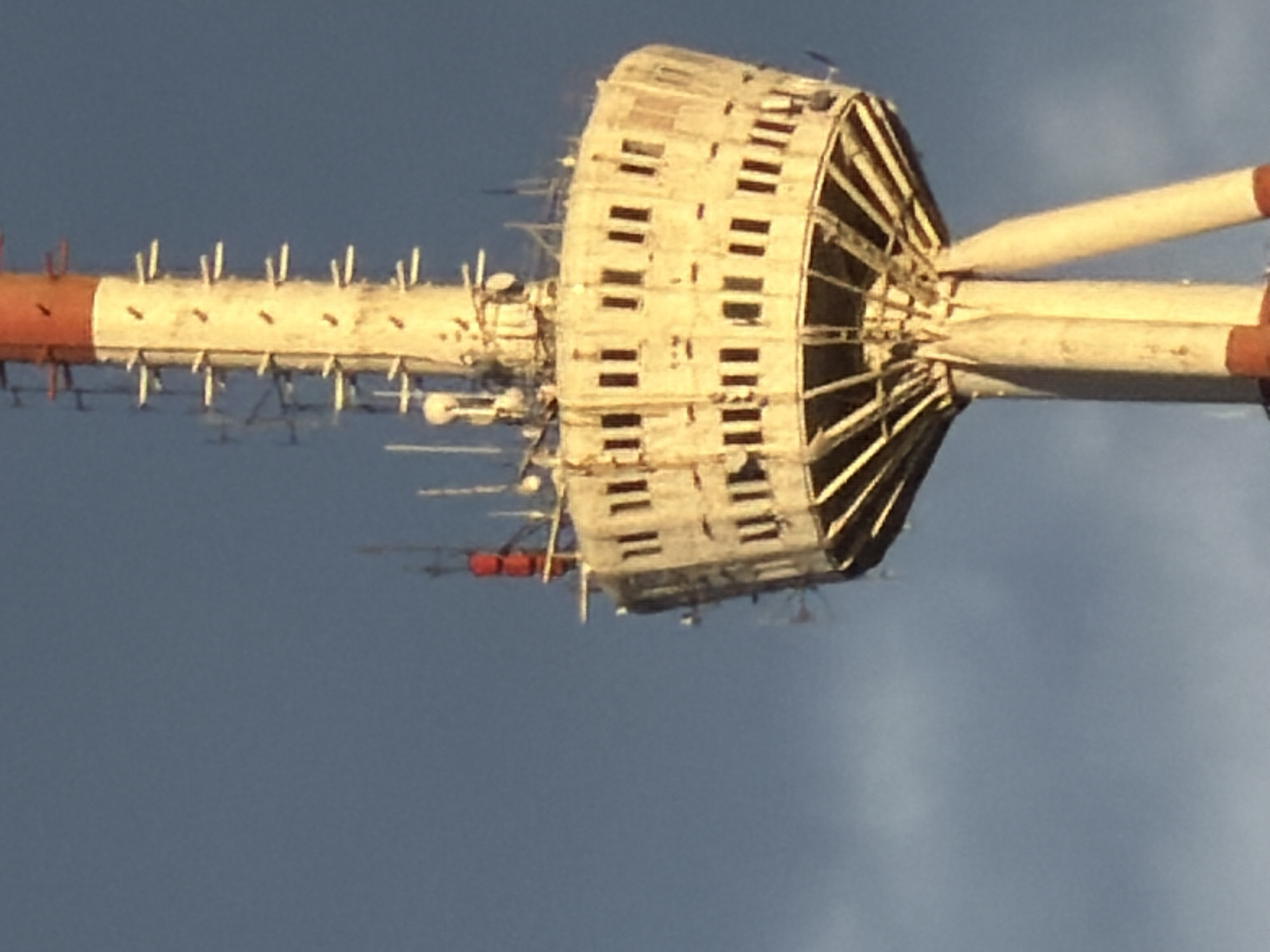 The image shows the television tower in Tbilisi, Georgia, shot from a low angle, looking upwards toward the sky. The tower is white and red, cylindrical in shape, with a round lattice structure near the top. There are various antennae and other attachments along the length of the tower. The sky is a light blue color.