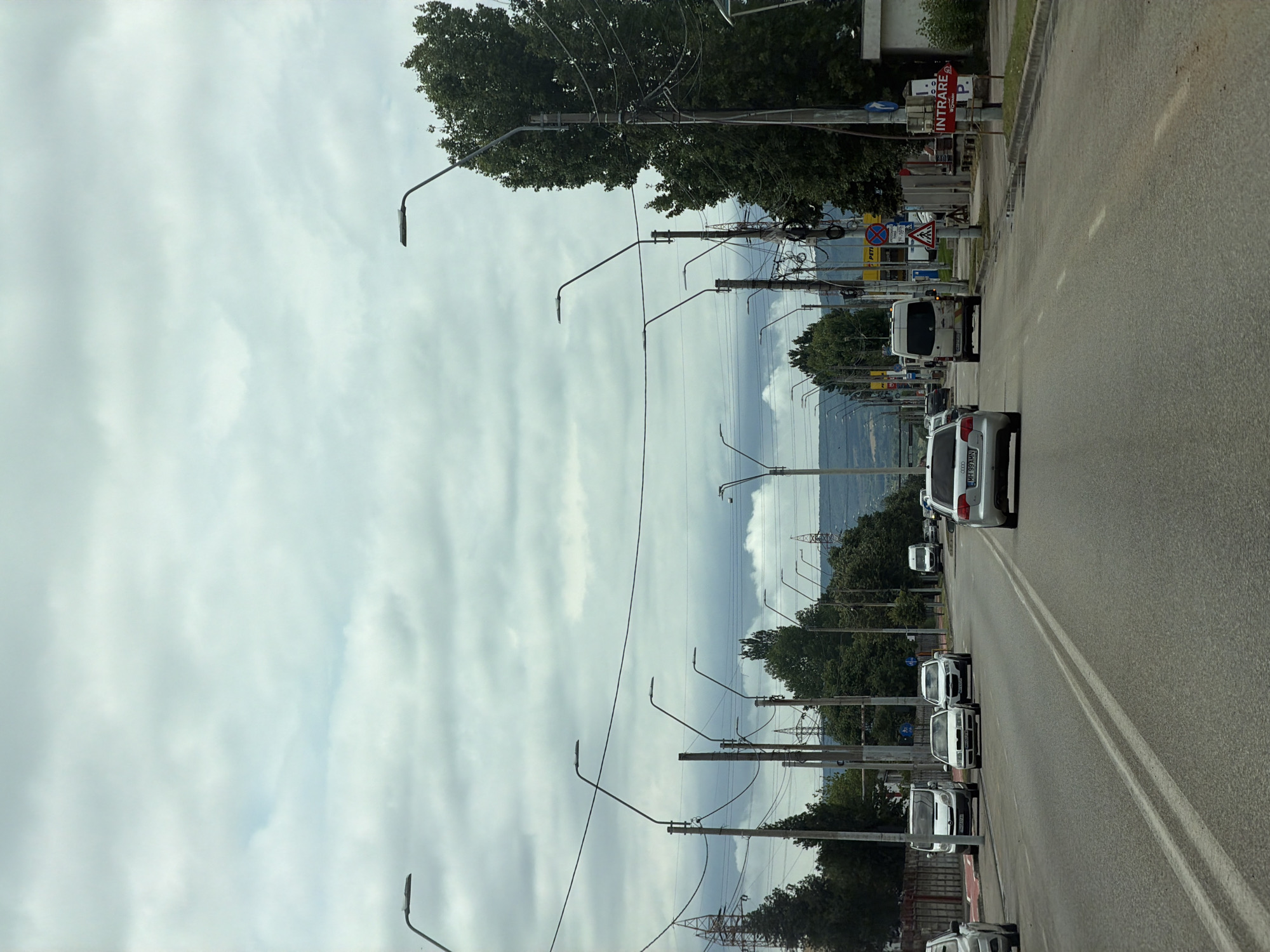 This media file captures a street scene in Drobeta-Turnu Severin, Romania, likely during daytime under an overcast sky. The image is taken from a slightly elevated perspective, looking down a two-lane road.
**Subjects:** The primary subjects include a long stretch of asphalt road with double solid white lines down the center, numerous utility poles fitted with streetlights and dense overhead wires, and a variety of vehicles. These vehicles consist of several white cars (sedans and what appears to be a van), some parked along the right side of the road, and others potentially in traffic. Lush green trees line both sides of the street, and in the distance, hills or mountains are visible, interspersed with large power pylons. The sky above is largely covered by diffuse clouds.
**Activity or event taking place:** The scene depicts typical daily activity on a street, with vehicles parked or moving.
**Notable details:** The weather is cloudy, contributing to a soft, even light across the scene. The utility poles are particularly prominent, with a multitude of wires crisscrossing overhead. On the right side of the road, several signs are visible. A red rectangular sign clearly reads "INTRARE P." (Romanian for "Entrance Parking"). Below it, a blue circular "no parking" sign is visible, along with a yellow sign that partially shows the text "PITII". A triangular warning sign for a pedestrian crossing is also present. The license plate "MH 99 MUN" is clearly visible on a white sedan in the foreground, and another plate starting "MH 02" is on a car further down, confirming the location in Mehedinți County, where Drobeta-Turnu Severin is situated. The overall impression is that of an active urban or suburban street.