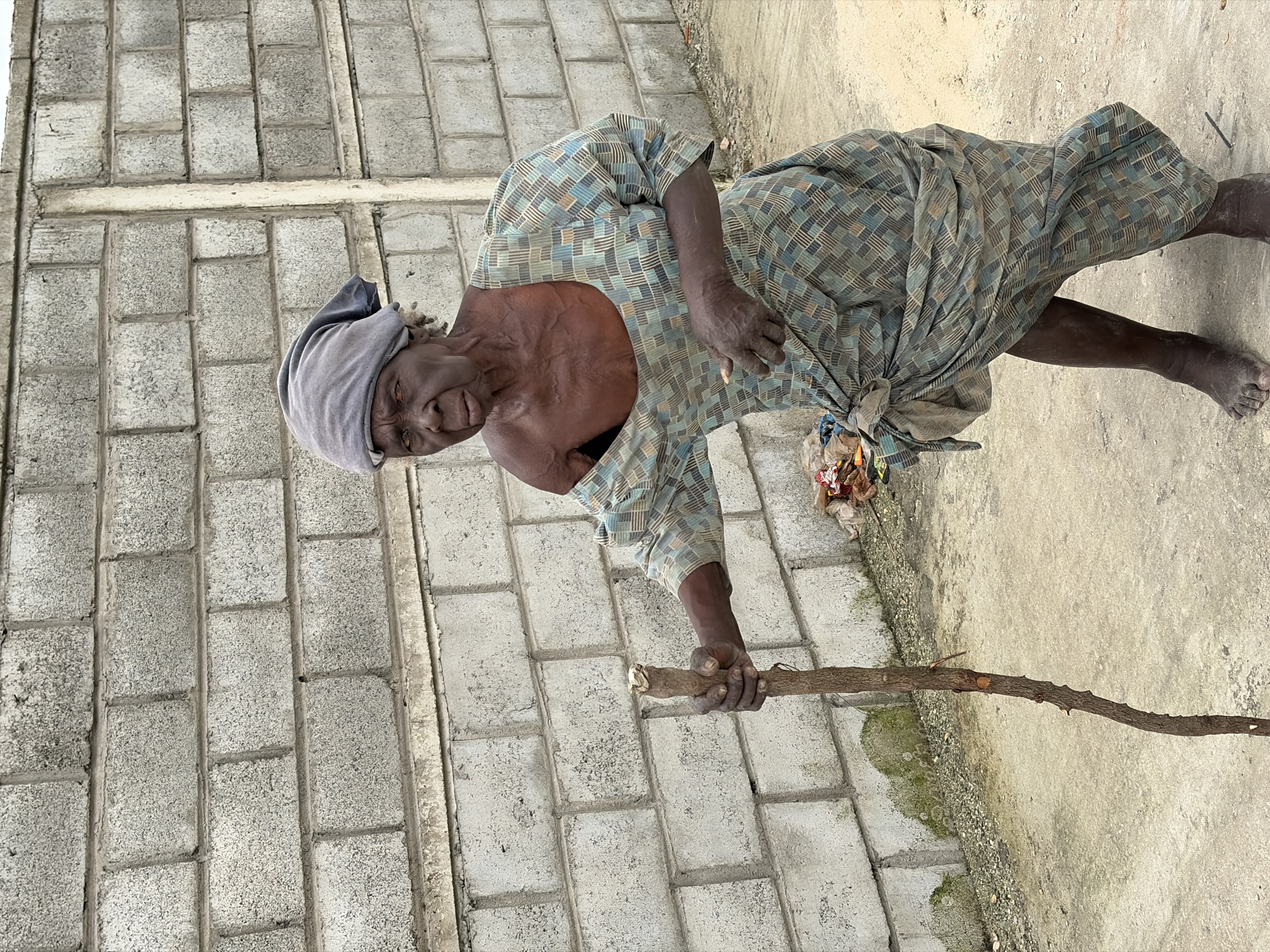 An elderly woman, with dark skin and wrinkled features, stands on a dirt path in Igboefon, Nigeria. She is wearing a light blue and green patterned dress and a gray headscarf. Her right hand grips a thick wooden walking stick. Her left hand is partially hidden by the fabric of her dress, which is cinched at her waist, forming a makeshift pouch. In the background, a wall constructed of grey concrete blocks stretches across the frame, with a vertical concrete post interrupting the pattern. The ground is a mixture of dirt and small gravel. The scene is lit by natural daylight.