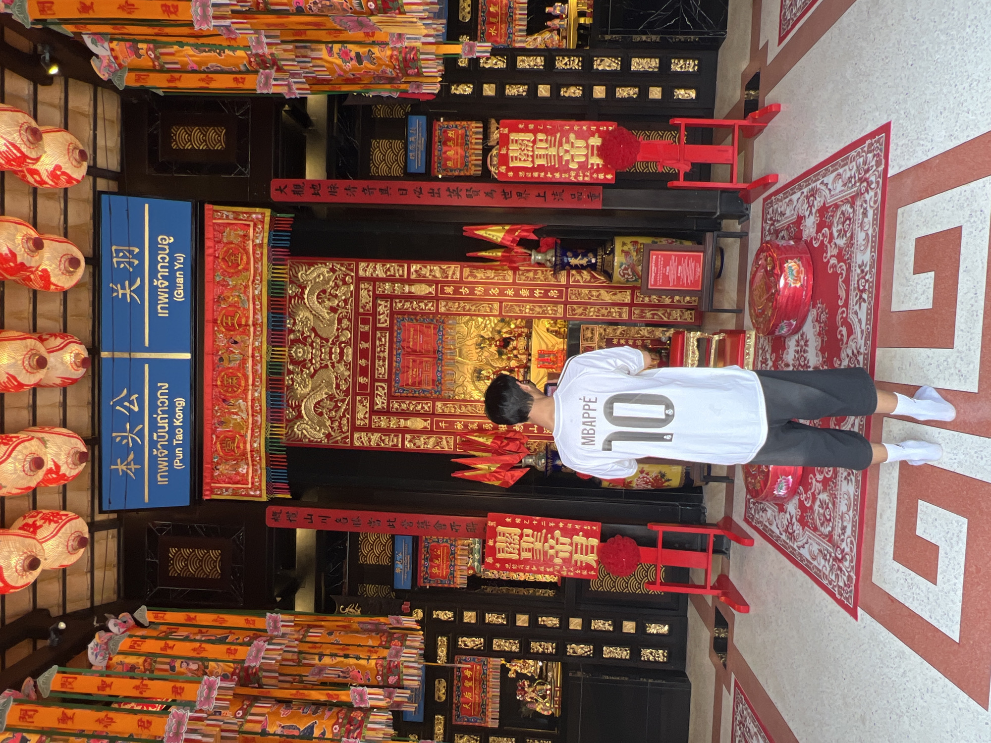 The image shows a person from behind, wearing a white t-shirt with the name "MBAPPÉ" and the number "10" on the back, along with black pants and white socks. They are standing in a temple or shrine, suggested by the ornate decorations, red and gold colors, and the presence of lanterns and scrolls. The scene appears to be outdoors or in a covered outdoor area, as indicated by the natural lighting and the open arrangement of elements.
The setting is richly decorated with traditional Chinese motifs, including dragon carvings and elaborate patterns on scrolls and banners. Several signs with Chinese and Thai text are visible, identifying deities and possibly providing information about the temple. Some of the visible text includes "關聖帝君" (Guān Shèng Dì Jūn, likely Guan Yu, a deified general), "เทพเจ้ากวนอู" (Guan Yu God) and "เทพเจ้ายืนถ่าวกง" (Pun Tao Kong God). The overall atmosphere seems to be one of reverence and cultural significance. The lighting suggests it could be daytime, though the exact time is unclear. The presence of multiple lanterns and ceremonial items points towards a place of worship or cultural celebration.
