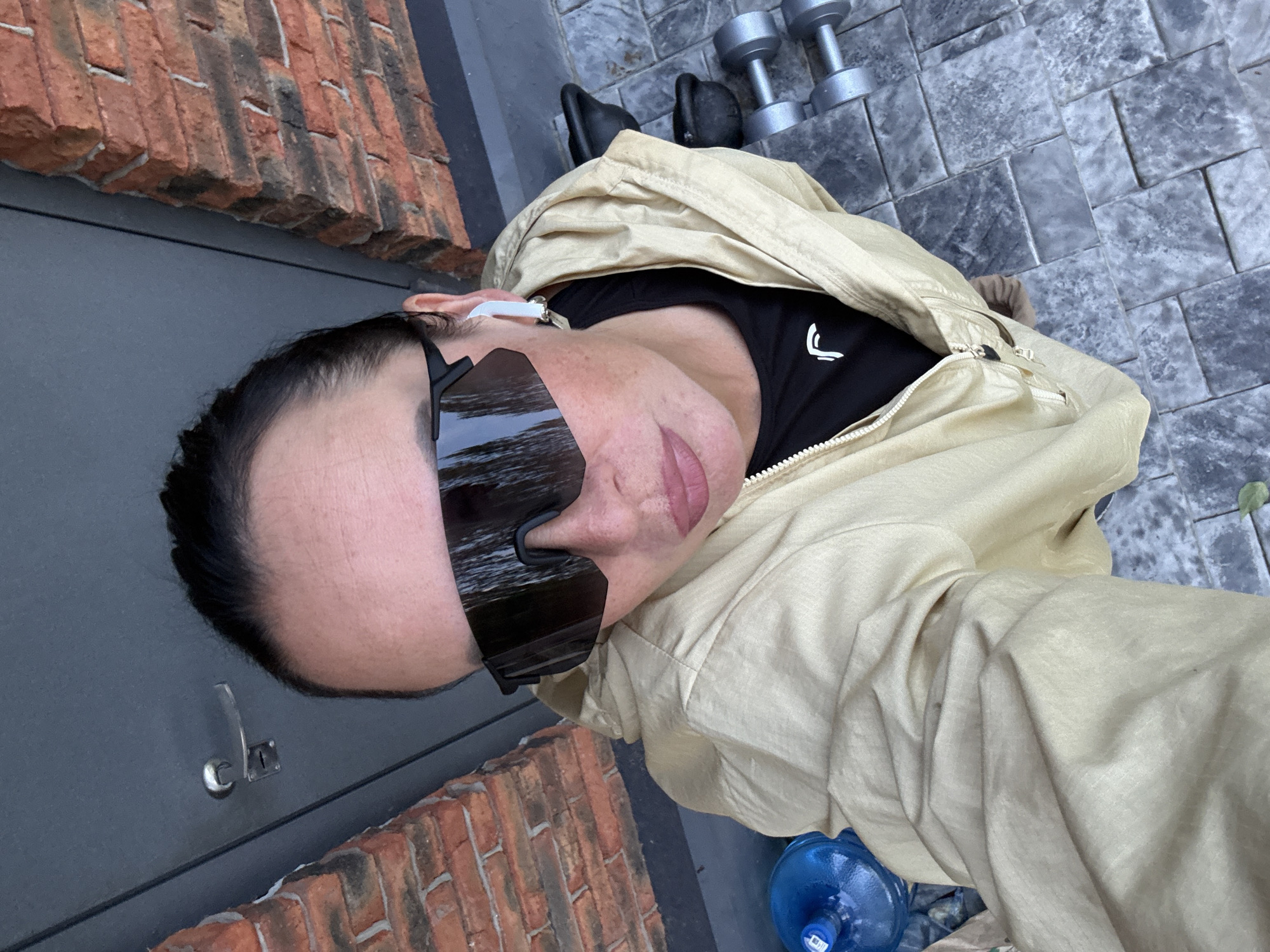 A woman, wearing sunglasses and a light-colored jacket, takes a selfie outdoors. She has dark hair pulled back and is wearing a black t-shirt with a white logo. White wireless earbuds are visible in her ears. The background shows a grey door with a handle, a brick wall, and a tiled floor. To her right are two kettlebells and two dumbbells. A large blue water bottle is to her left. The scene appears to be located outside a building in Chaikovsky, Russia.
