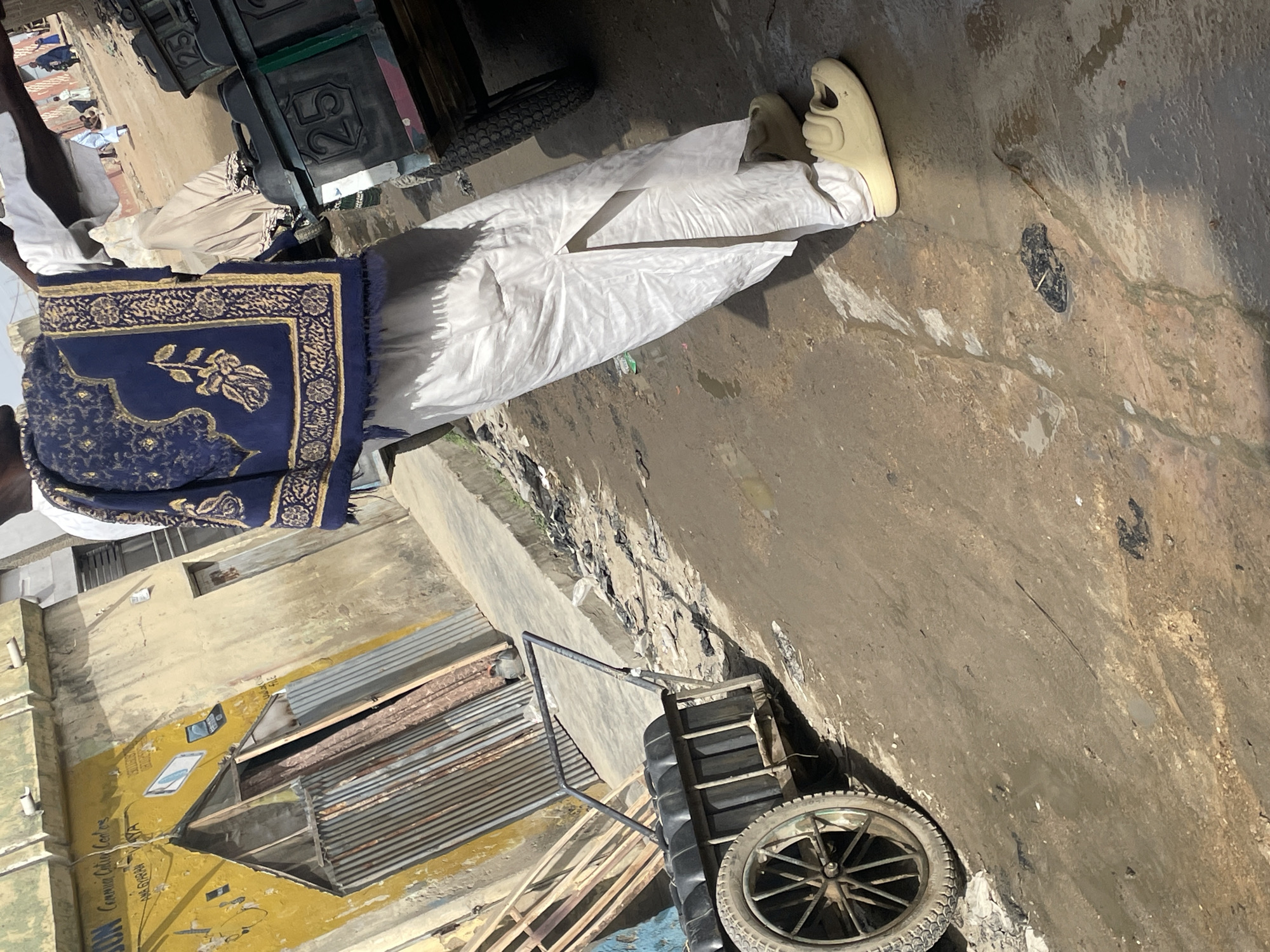 A man in traditional attire is seen walking on a street in an urban setting. The man is wearing a white outfit with a blue and gold patterned robe draped over his shoulder. He is wearing light-colored slip-on shoes. He is walking past a handcart with a single wheel and a number '25' on it. The street appears to be somewhat dirty, with possible signs of standing water or wet patches. In the background, there is a building with faded yellow paint and a corrugated metal roof. The location is Unknown, Unknown.