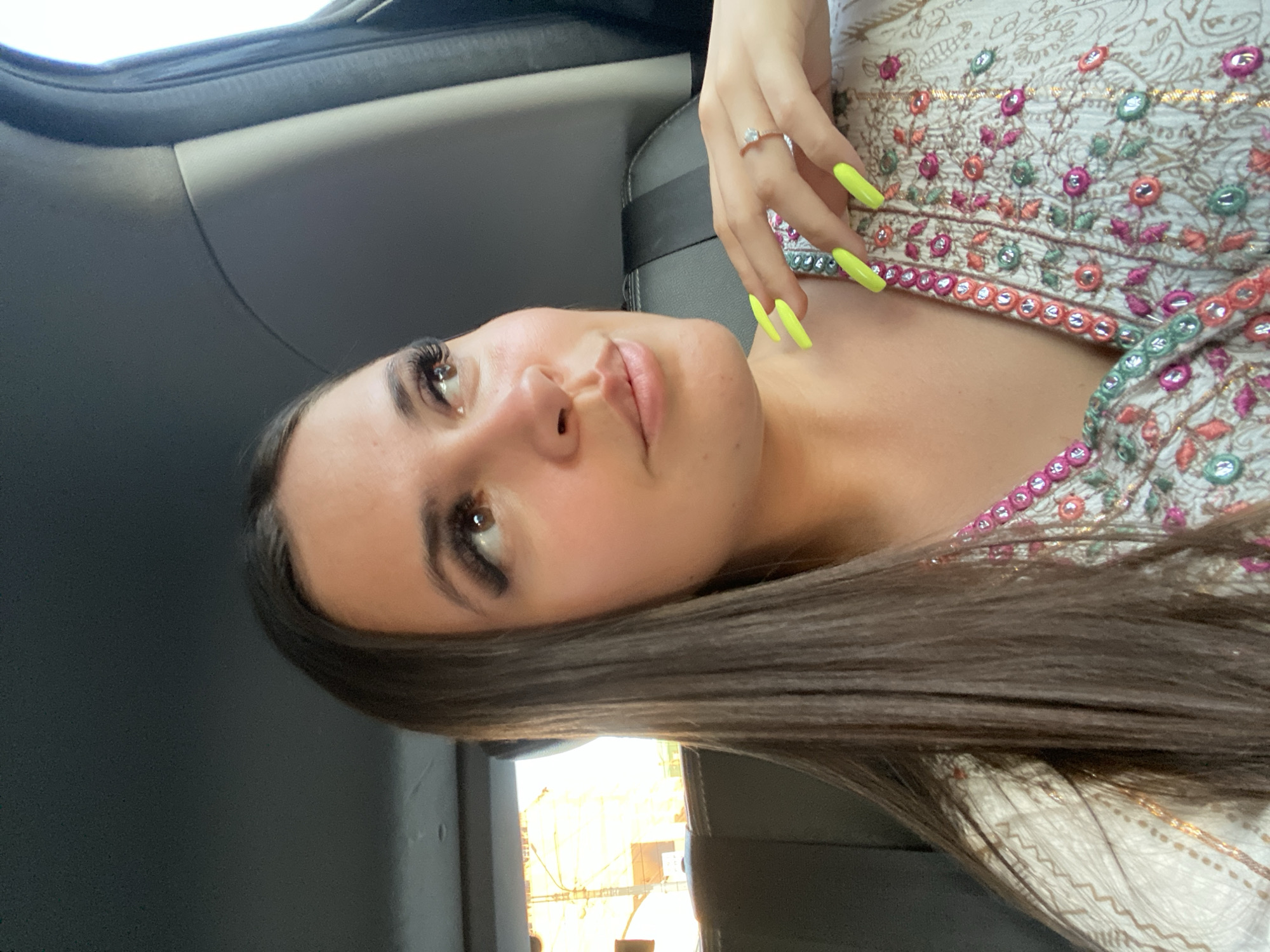 A young woman with long, dark hair and striking makeup, including prominent eyelashes and bright neon yellow fingernails, is pictured in a car. She is wearing a white top adorned with colorful embroidery and what appears to be a diamond ring on her left ring finger. The woman's gaze is directed slightly away from the camera, with a soft, contemplative expression. The interior of the car, with its dark gray upholstery, is visible, suggesting the woman is a passenger. The lighting indicates it is daytime, and the outdoor environment visible through the car window appears to be urban, possibly an industrial area given the distant structures.