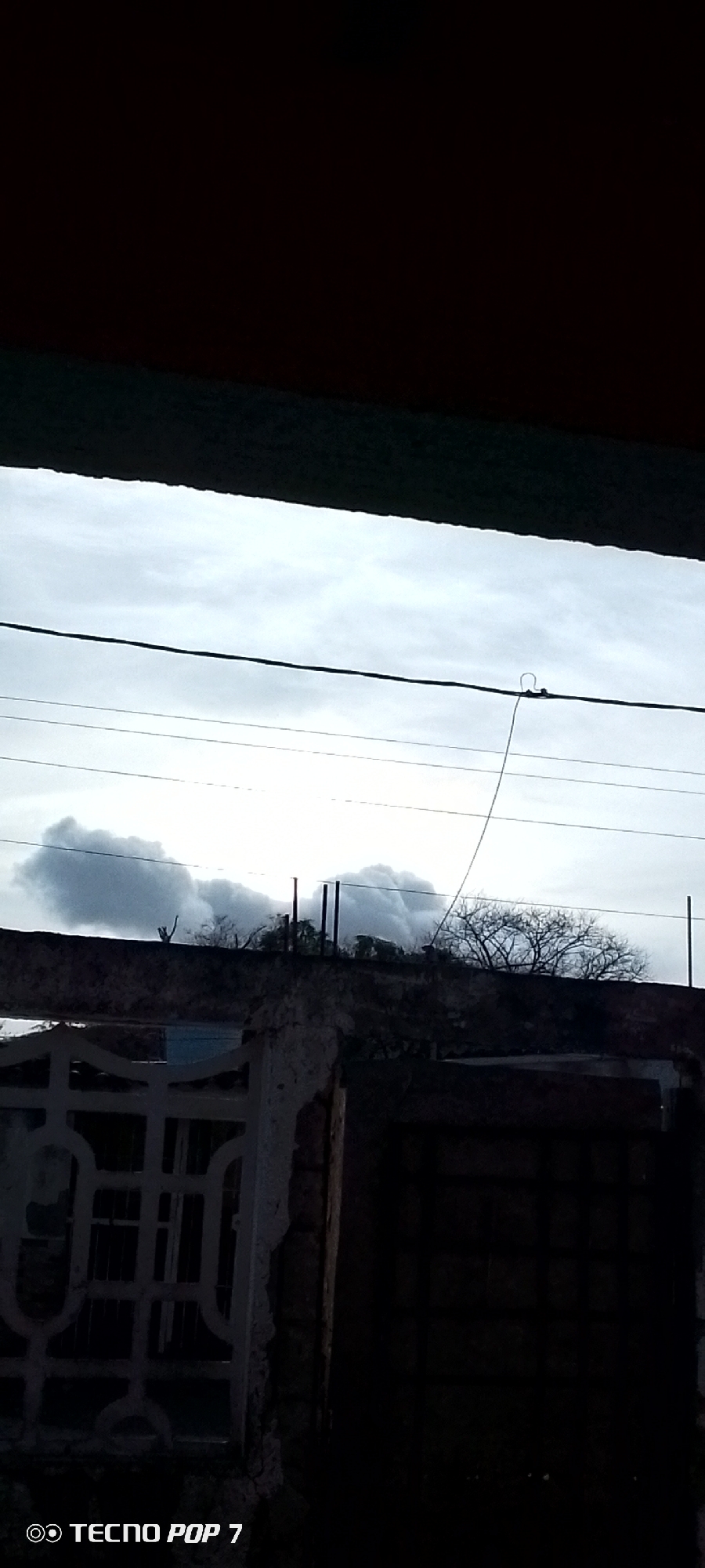 A view of Altagracia, Venezuela, looking out from a dark space with a partially visible, ornate white gate and a dilapidated brick structure. Above these, the overcast sky is seen with power lines running across the frame. In the distance, trees and what appears to be smoke or clouds are visible.