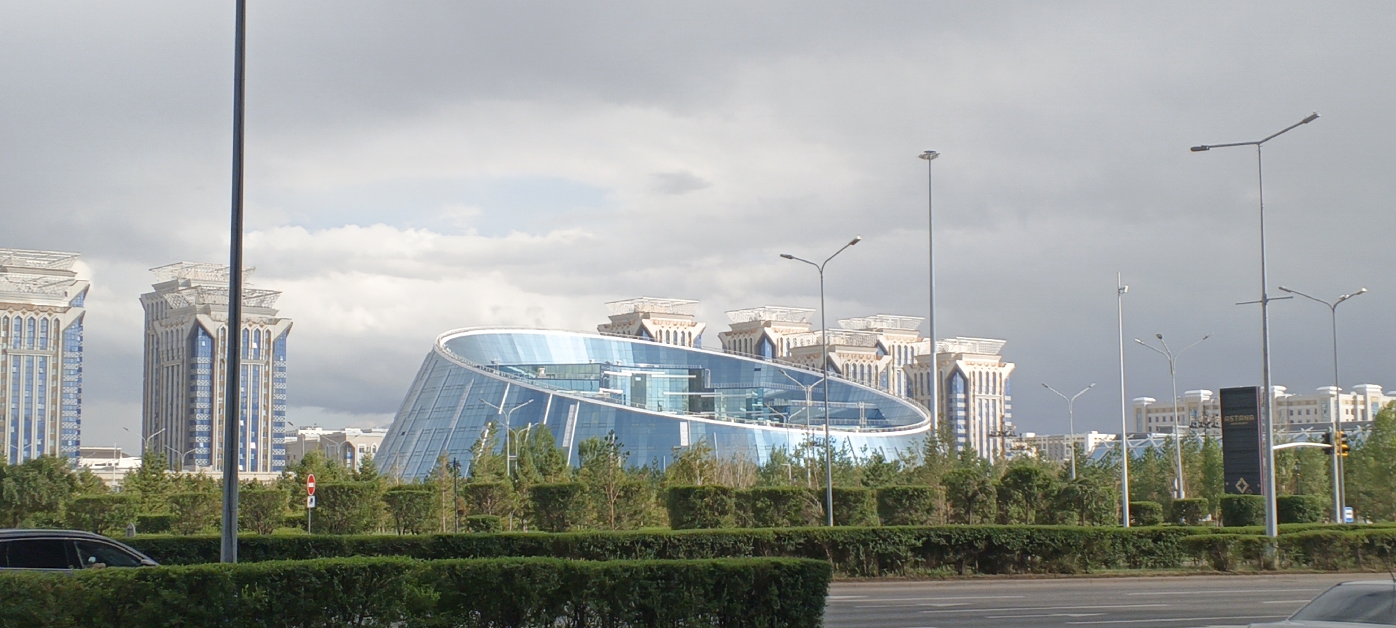 A modern cityscape in Astana, Kazakhstan, features a large, curved glass building with a futuristic design. The building's facade reflects the cloudy sky. In the foreground, there are several streetlights, a tall vertical pole, and a dark rectangular sign with the word "ASTANA" visible. A low-lying green hedge runs horizontally across the bottom of the frame, separating the street from the buildings and greenery behind it. Cars are partially visible on the road. In the background, several tall, ornate buildings with pointed roofs and blue-tinted windows stand alongside the glass structure. Lush green trees and manicured bushes fill the mid-ground. The sky is overcast with varying shades of gray and white clouds.