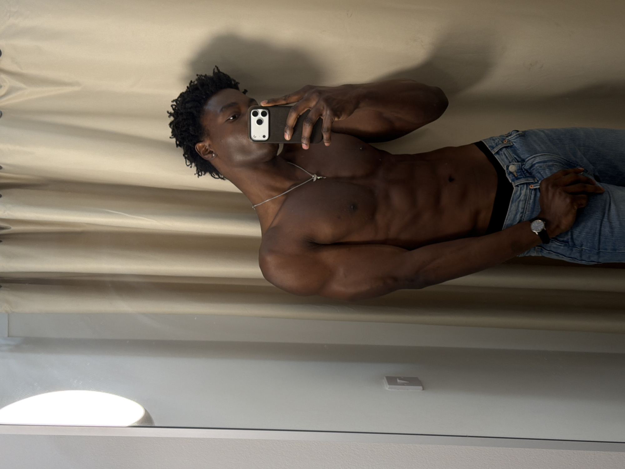 A tanned-skinned male is taking a photograph of himself in a mirror. He is wearing jeans, a silver necklace, and a watch on his left wrist. His torso is bare. The background consists of a tan-colored curtain and a white wall. A light source is visible in the lower-left corner. There is no information to suggest a location in Manchester, United Kingdom.