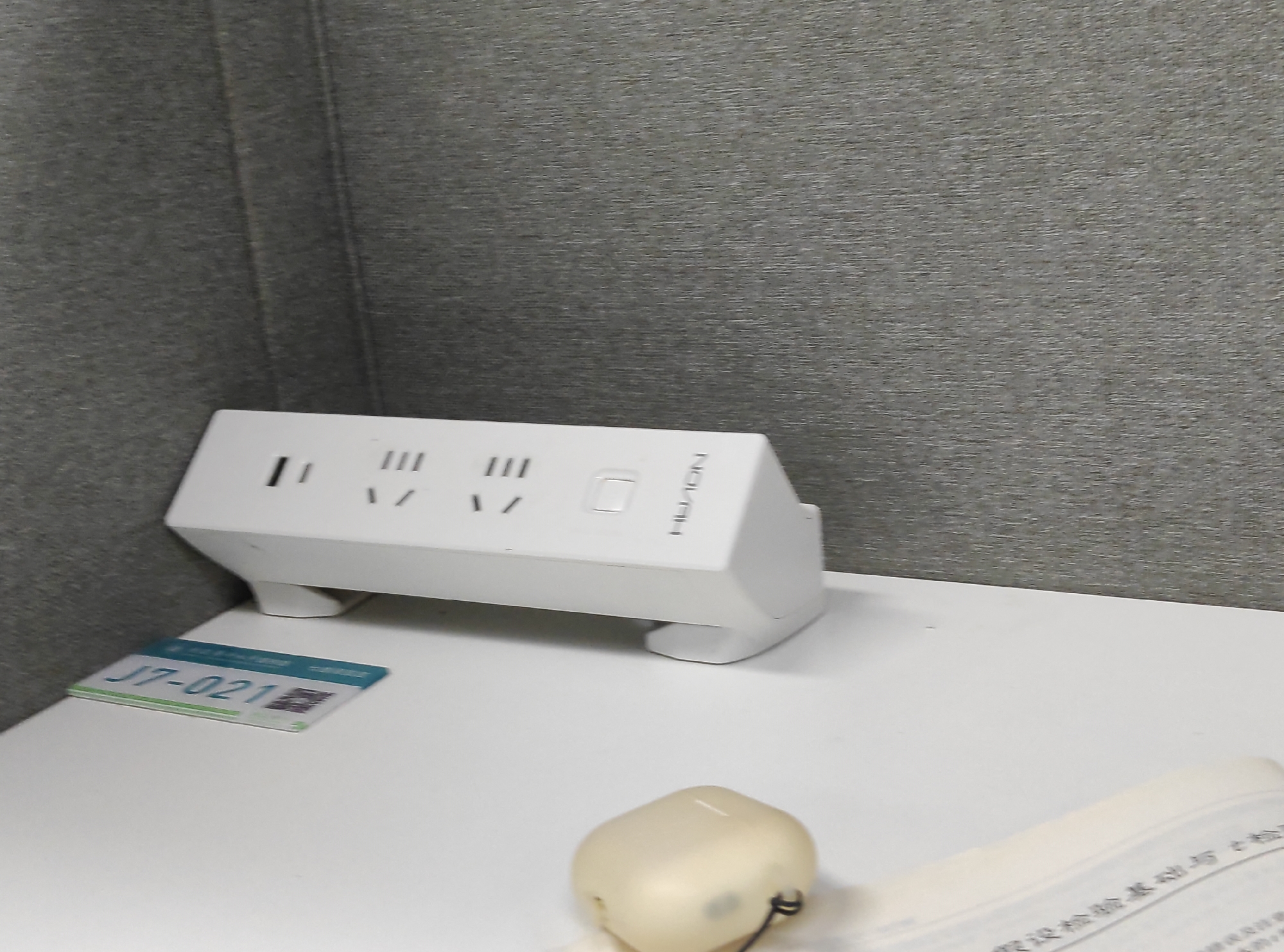 An overhead shot captures a white power strip with three sets of outlets and a silver button on a white desk. A label with green text and a QR code is placed to the left of the power strip. In the foreground, a light-colored, rounded object with a black cord is on the desk. The background features a textured gray wall.