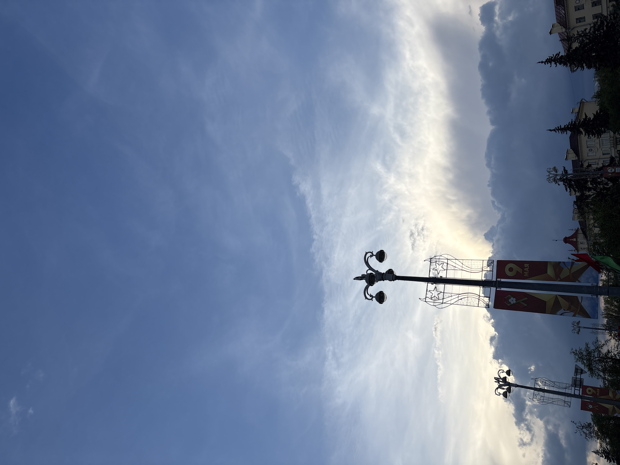 The image depicts a sky scene with a combination of clear blue and partly cloudy sections. In the foreground, a dark-colored ornate lamppost is visible, along with banners commemorating the 9th of May. Adjacent to the lamppost, there are buildings. The photo appears to be taken in Minsk, Belarus.