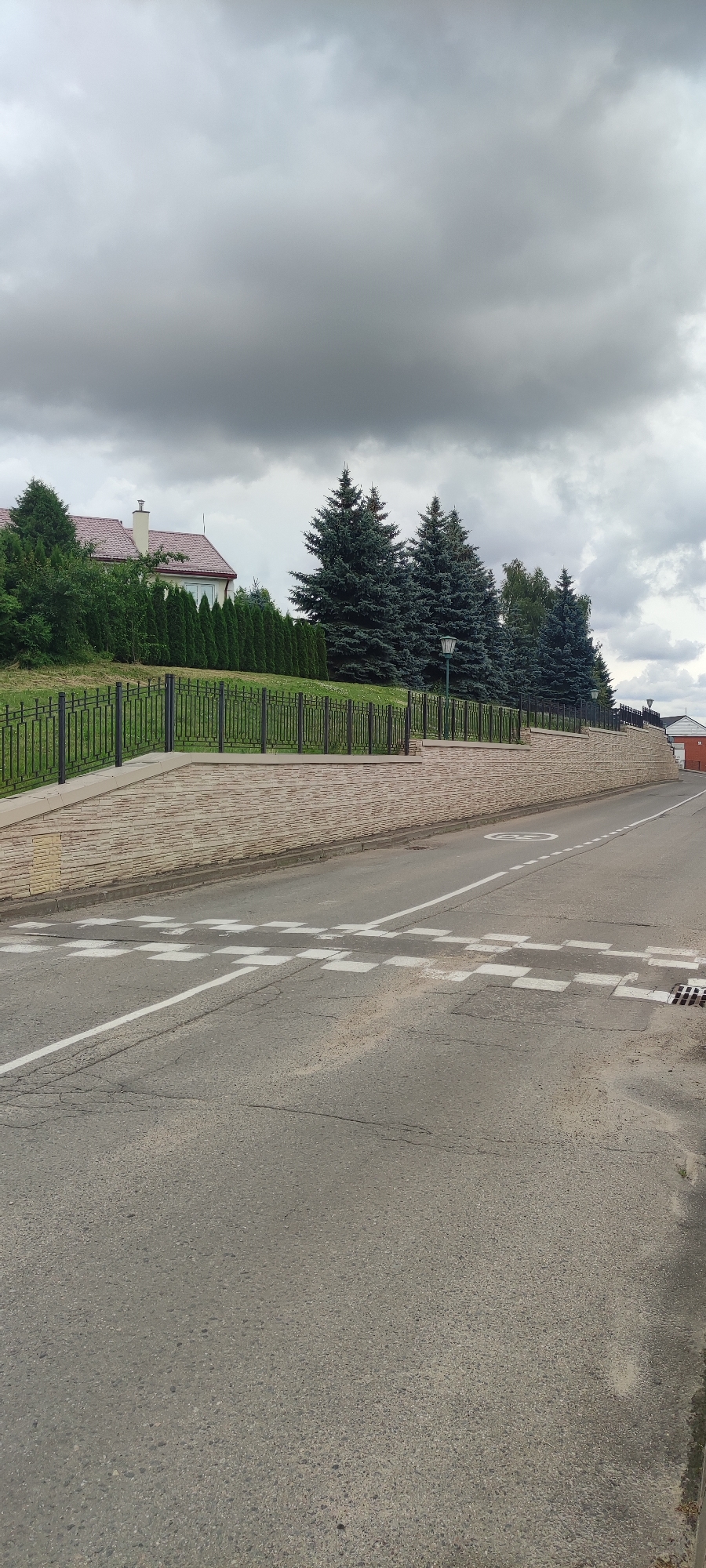 The image depicts a street scene in Rosinka, Russia, on a cloudy day. A two-lane asphalt road runs diagonally from the bottom right to the upper left of the frame. White lines mark the lanes and a pedestrian crossing with checkered white markings is visible on the road.
To the left of the road, a retaining wall constructed of light-colored, textured blocks rises from the sidewalk. Above the wall, a black metal fence encloses a grassy area. In the background, several tall evergreen trees stand in front of a building with a red-tiled roof and a white chimney. A row of manicured green bushes lines the property in front of the house. Another section of the retaining wall, lower in height, extends further into the background on the right side of the road, where a small portion of a red brick building is visible. The sky is filled with heavy, gray clouds. There are no people visible in the image.