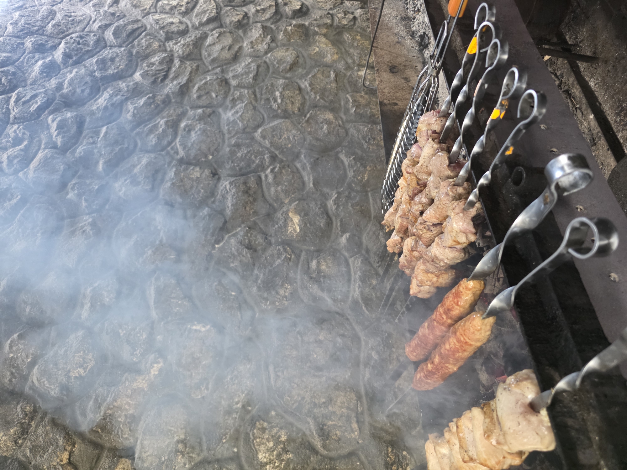 The image shows meat cooking on skewers over coals. The meat appears to be pork, with some pieces on skewers resembling kebabs and others appearing to be larger cuts of meat. Two sausages are also visible on skewers, and they are a reddish-brown color. The meat is positioned over a grate which is situated above a bed of glowing coals, indicating a cooking process. Smoke is rising from the cooking meat and dissipating into the air. The background consists of a stone wall, which has a textured, irregular pattern. The scene appears to be outdoors, possibly in a rural setting given the stone wall and the open-air cooking setup. The lighting suggests it is daytime, but the specific time is not discernible. The overall impression is one of food preparation, likely for a meal or gathering. The location context provided is Pidhorodne, Ukraine. There is no visible text in the image.