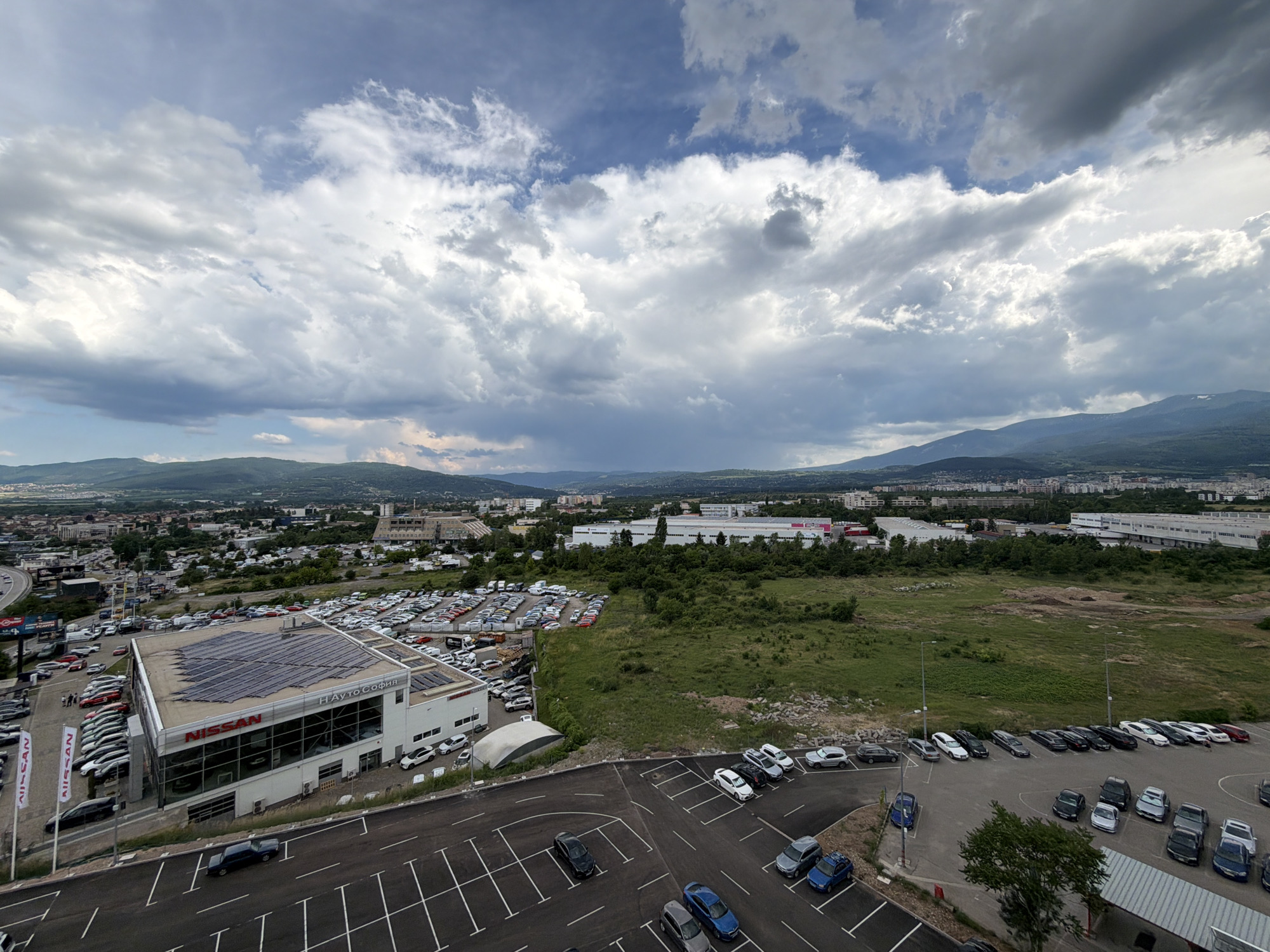 An aerial, wide-angle view captures a car dealership in Sofia, Bulgaria, on a cloudy day. The dealership, identified by the "NISSAN" logo on its building, is a prominent structure with a large parking lot filled with numerous vehicles. Adjacent to the dealership, a vast open field stretches towards a city skyline dotted with buildings. In the background, rolling hills and mountains, some with snow-capped peaks, form the horizon under a dramatic sky filled with cumulonimbus clouds. The overall scene depicts an urban landscape interacting with natural surroundings.