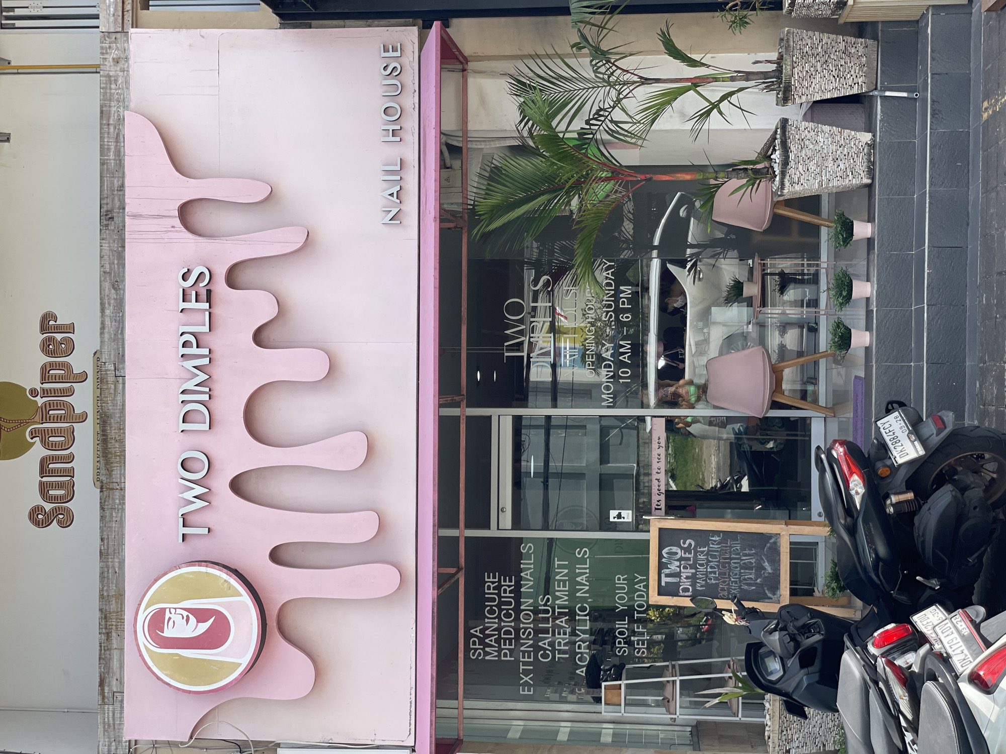 This is the exterior of the Two Dimples Nail House in Kuta Selatan, Indonesia. It is a pink-themed nail salon with a distinct logo and signage. The sign prominently displays "TWO DIMPLES NAIL HOUSE." The front of the salon is adorned with plants in decorative pots. Services advertised include spa treatments, manicures, pedicures, extension nails, callus treatments, and acrylic nails. The storefront has glass doors and windows, and a chalkboard sign advertises the salon's services. Several motorbikes are parked in front of the establishment. The adjacent business is named "Sandpiper."