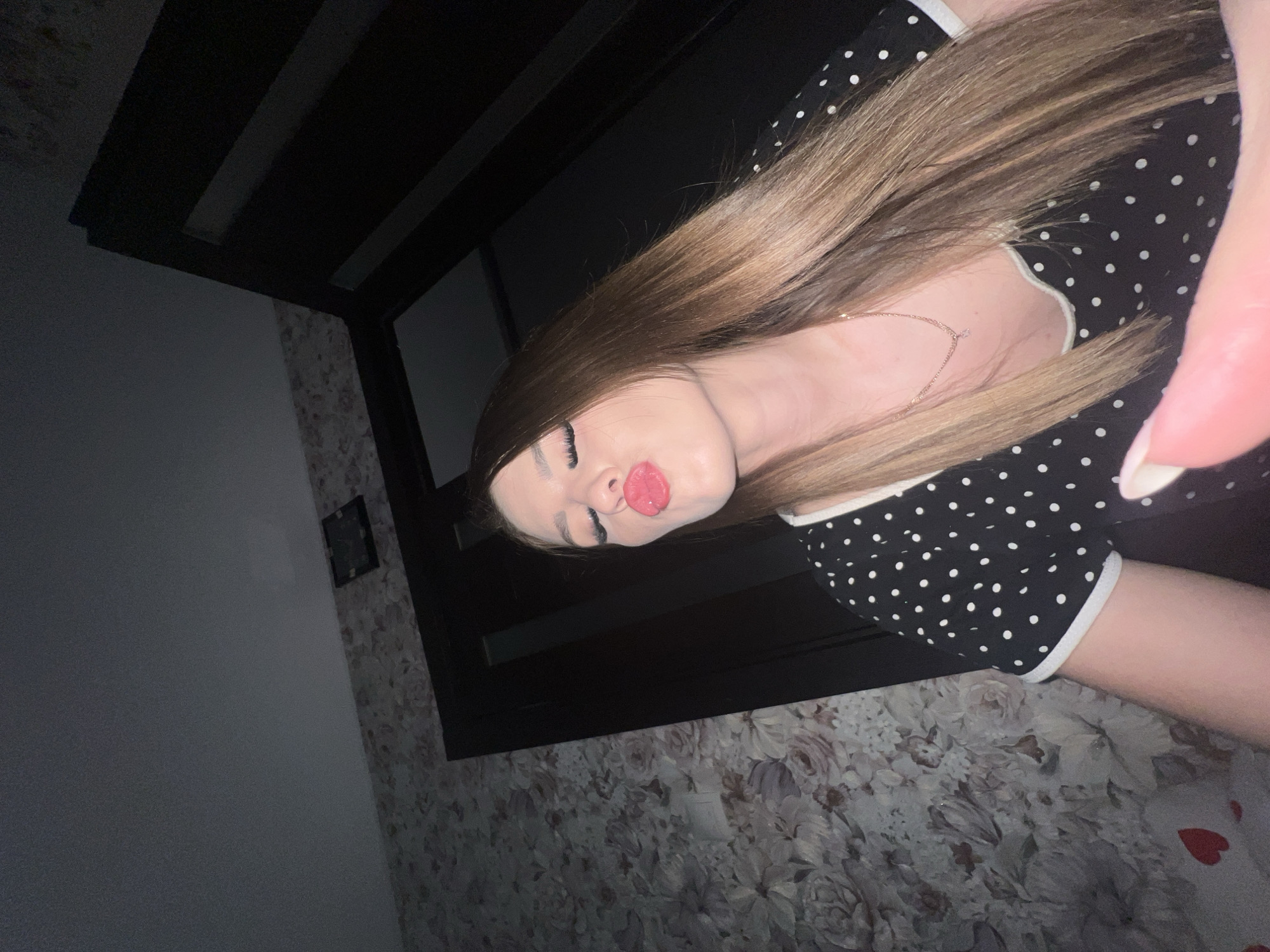 A young woman with long, straight brown hair and bright red lipstick is captured in a selfie. She is wearing a black polka dot dress with white trim and a delicate gold necklace. Her eyes are closed, and her lips are pursed in a kissing gesture. The background consists of a wall with floral wallpaper and a dark doorway. The lighting suggests an indoor setting, possibly a home, at night. There are no discernable elements in the image that would indicate a specific location within the city of Noviy, Russia.