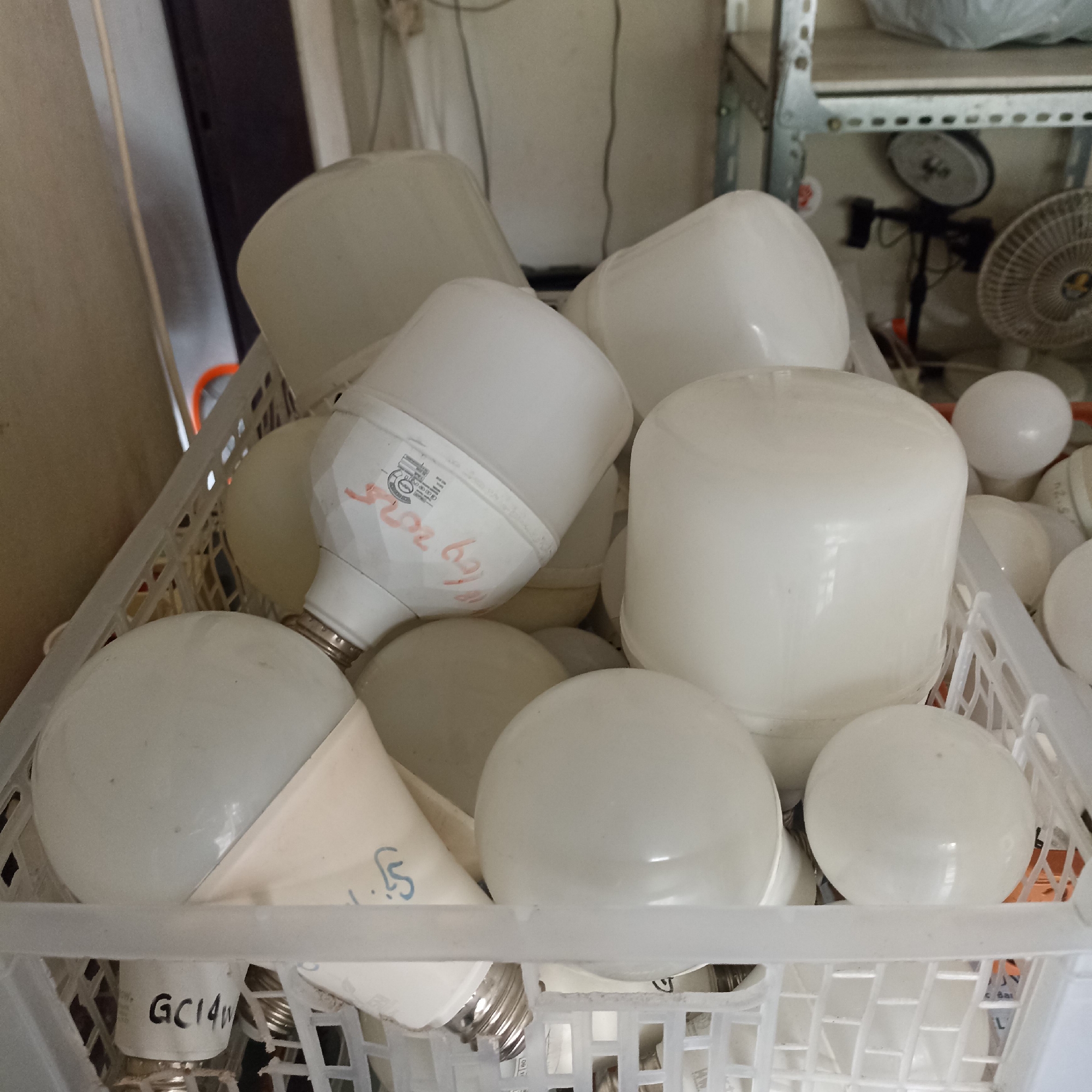 A close-up, angled shot shows a white plastic basket filled with white LED light bulbs. The basket is overflowing with the bulbs, some of which are stacked on top of each other. The bulbs vary in shape and size, with some being more cylindrical and others more spherical. Some of the bulbs have labels or writing on them, but they are largely obscured by the other bulbs or are too blurry to read. In the background, a metal shelf unit is visible, along with other objects that are out of focus, including what appears to be a fan. The overall impression is one of storage or perhaps a retail setting. The location is identified as a storage area in Magelang, Indonesia.