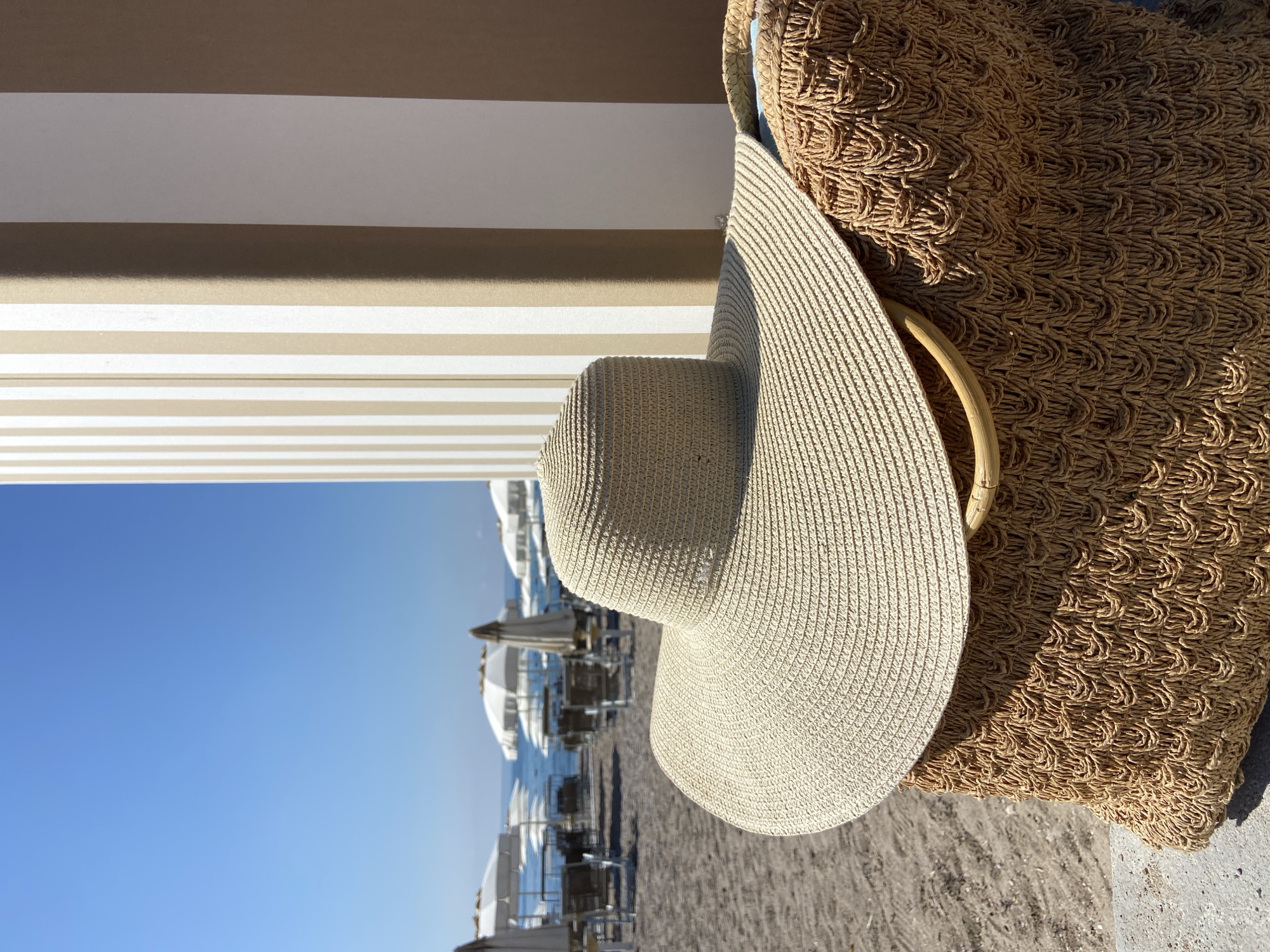 In Prijevor, Montenegro, a wide-brimmed straw hat rests on top of a woven straw bag. The bag has a textured pattern and a light-colored wooden handle. Behind the hat and bag, a sandy beach is visible with rows of empty lounge chairs and white umbrellas. The calm sea stretches to the horizon under a clear blue sky. To the right, a structure with alternating vertical stripes of beige and white stands partially in view. The scene depicts a peaceful beach setting, likely before or after peak visitor hours.