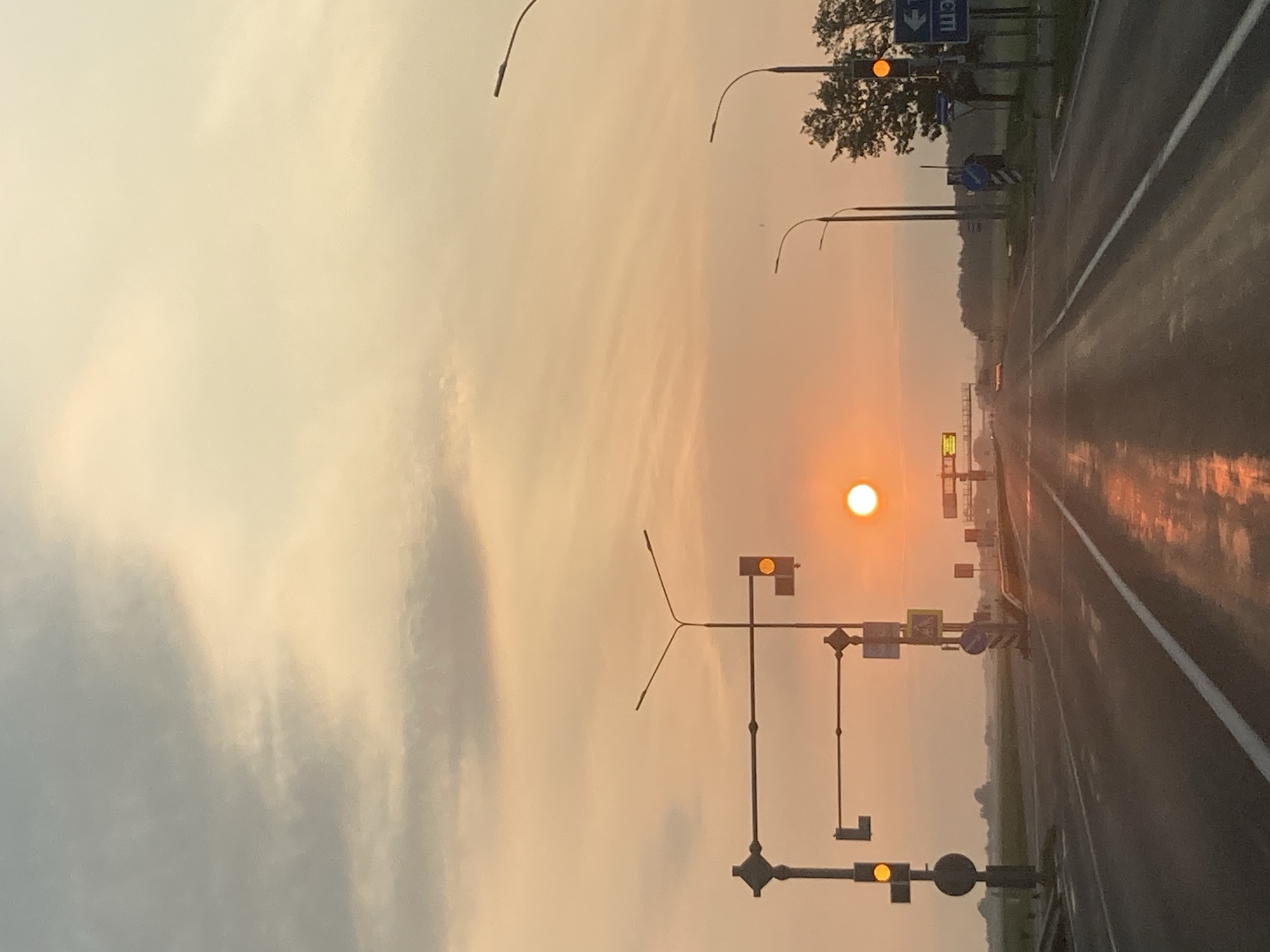 The image depicts a roadside scene at sunrise or sunset, with the sun visible as a bright orb in a hazy, orange-toned sky. A two-lane asphalt road stretches into the distance, wet and reflective, suggesting recent rain. Traffic signals with illuminated yellow lights are present on both sides of the road. Road signs, including directional signs and a pedestrian crossing sign, are also visible. Streetlights line the road. The landscape appears rural, with fields and a treeline in the background. The location is identified as Тэльмінскі сельскі Савет, Belarus. No people are visible in the image.