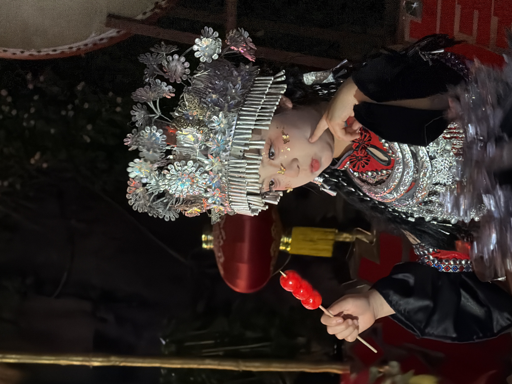 In a dimly lit, outdoor setting, likely Yuzhong District, China, a young child, possibly of ethnic minority descent, is the central focus. They are adorned in a traditional, elaborate silver headdress, intricately detailed with floral motifs and hanging ornaments. The child wears a black garment with red and silver accents on the collar and chest, a silver collar piece, and shiny embellishments on the sleeves and hem. Their face is lightly made up with glitter and gold butterfly-shaped accents on the cheeks and forehead, and a touch of red lipstick.
The child is holding a skewer with three red, glossy candies (likely tanghulu) in their left hand, bringing it close to their mouth with their right index finger touching their cheek. Their expression is one of mild curiosity or contemplation.
In the background, a large, red, silk lantern hangs, illuminated from within, and a yellow tassel is visible beneath it. Part of a large drum with a patterned rim is visible to the right, and a bamboo pole stands to the left. The overall ambiance suggests a festive or cultural event at dusk or night.