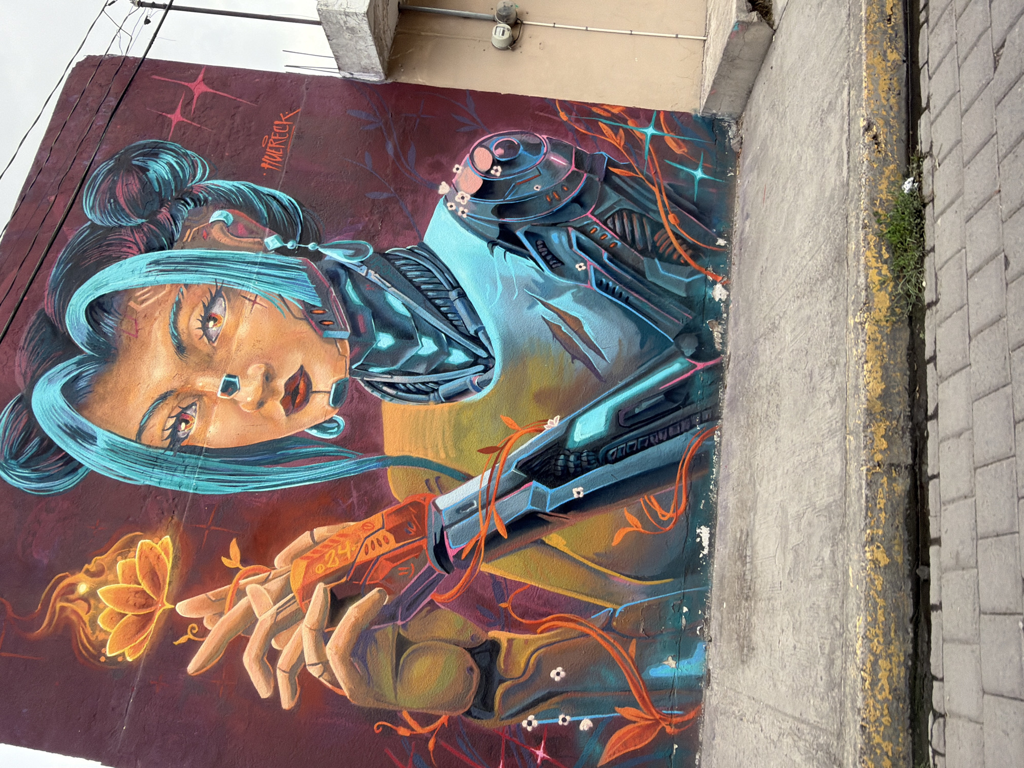 The image shows a mural on a wall in San Andrés Cholula, Mexico. The mural depicts a stylized woman with blue hair in a bun and mechanical augmentations. She wears what appears to be robotic armor on her upper body. One hand appears organic, the other robotic. She is depicted holding a glowing orange flower. The background is a deep red-purple color. The wall is next to a sidewalk paved with gray bricks. A curb with yellow paint runs along the edge of the sidewalk. A building is visible above the mural. Electrical wires run above the building.