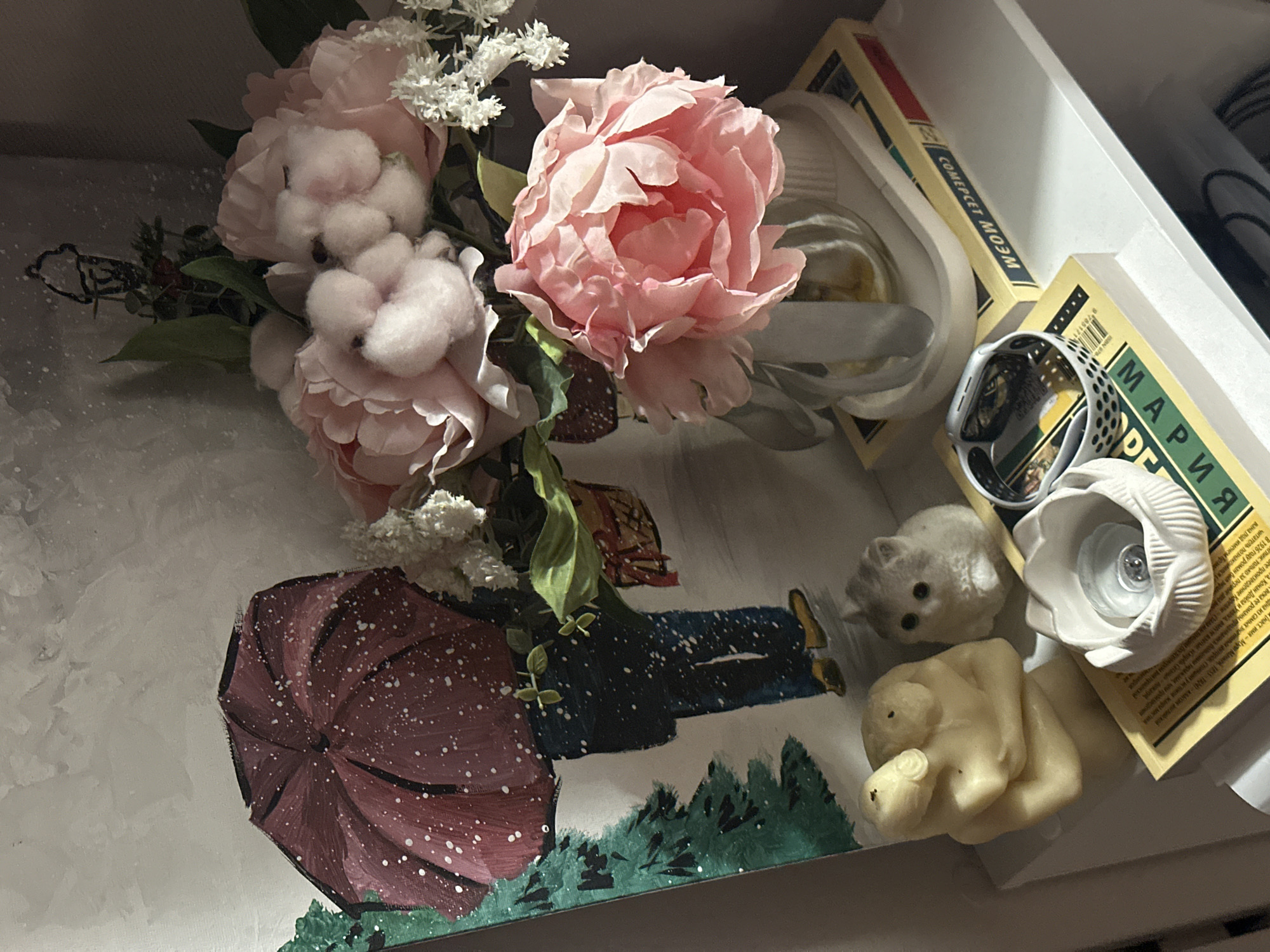 A still life composition resting in a container, possibly a drawer or box.
The scene is dominated by a cluster of pink peony-like flowers with white accents, intermingled with cotton blossoms and green leaves. Adjacent to the flowers is a depiction of a painted scene on a flat surface, showing an individual in dark clothing, with yellow shoes, under a red umbrella with white dots, over a background of white and green grass. To the right, there are small figurines including a hedgehog-like figure and a yellow sculpture of intertwined human figures. Various items are stacked beside them, including books or boxes with Russian text and branding. One of the boxes reads "COMEPCET WEOW" (Comercet Wow). Another label reads "MAPUA DE". In the background, a white object, possibly a container, is visible.