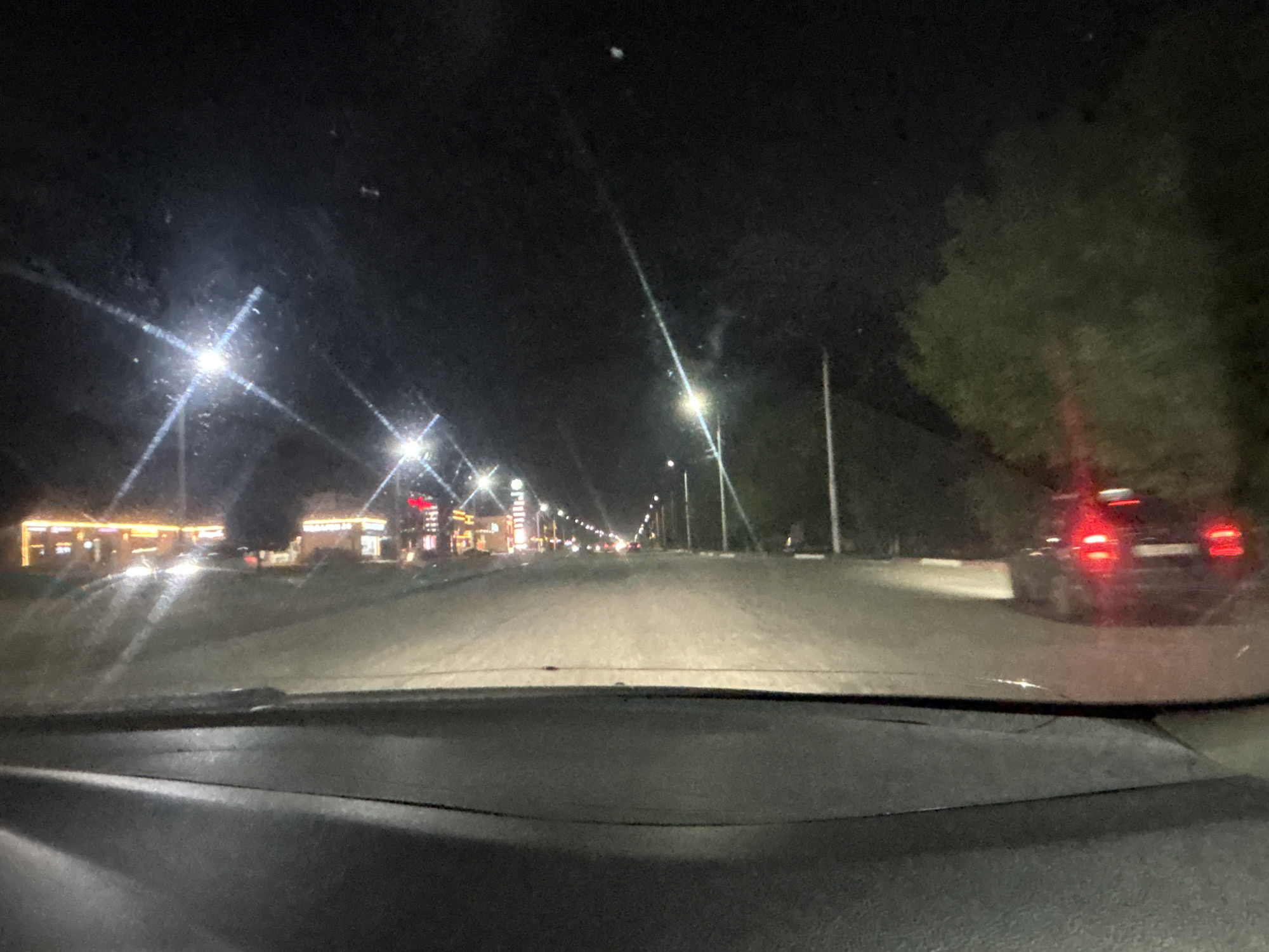 The image depicts a nighttime view from inside a vehicle, likely in the city of Grozny, Russia, as evidenced by the presence of street lights, buildings with illuminated signs, and a road. The scene is characterized by darkness punctuated by artificial light sources. The road ahead is well-lit by street lamps and the headlights of approaching vehicles. On the left side of the road, several buildings with glowing signs are visible, suggesting a commercial or entertainment district. One building has a prominent red sign. On the right side of the road, a dark-colored car with illuminated red taillights is partially visible, suggesting it is either parked or moving away. The overall environment appears to be a suburban or urban setting with trees lining the right side of the road. The interior of the vehicle, specifically the dashboard, is in the foreground, partially obscuring the view. The image has a somewhat grainy quality, typical of nighttime photography.