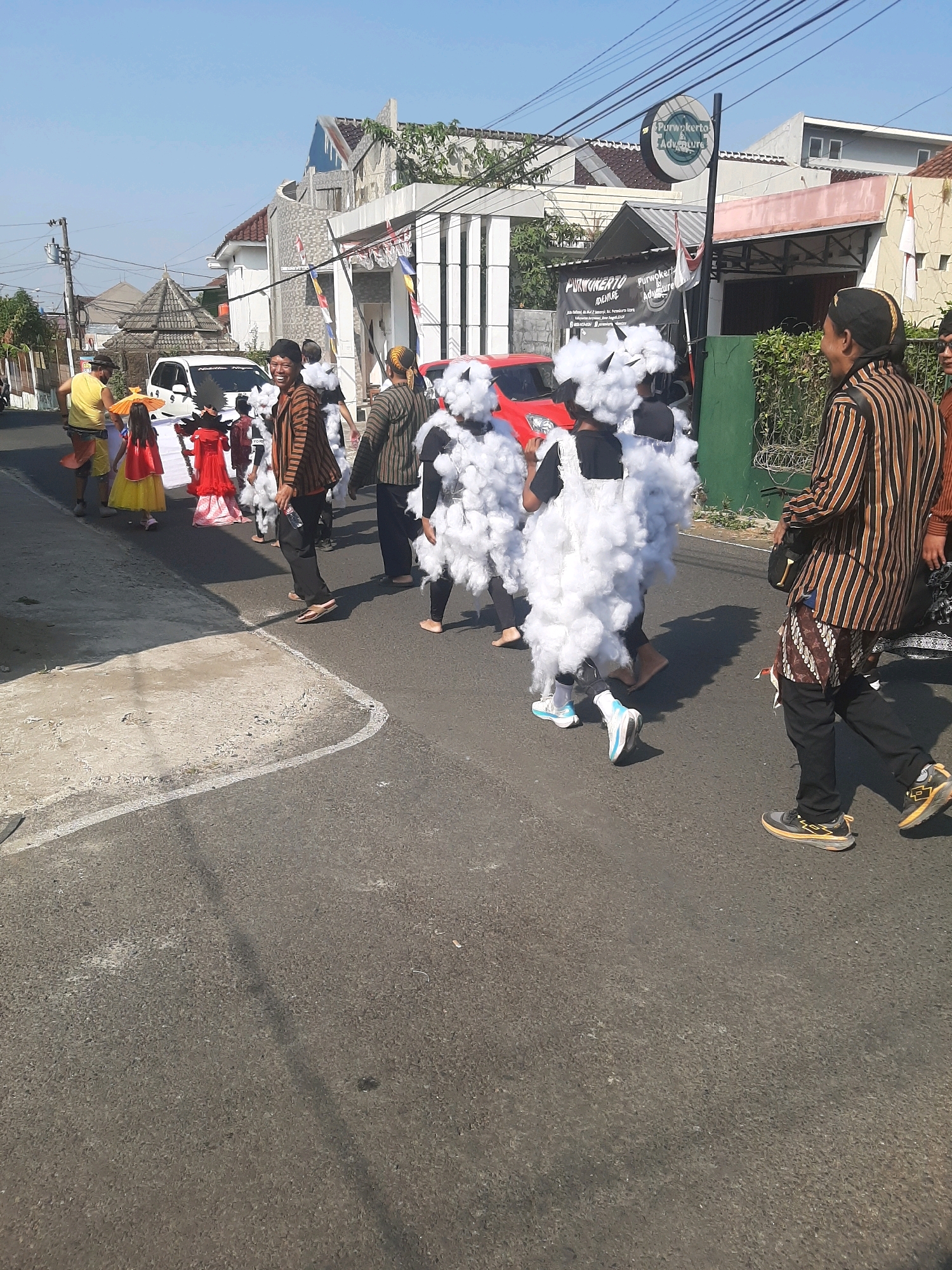 In Sumampir, Indonesia, a street parade is in progress on a paved road under a clear blue sky. The parade features several individuals, including two children dressed in large, white, cloud-like costumes with fluffy cotton material. One child wears a black dress under the costume, while the other wears a white undershirt and black pants. They are both walking forward, with one child looking towards the camera and smiling.
To their left, a group of people are also participating in the parade. One person in a yellow shirt and red skirt is holding a yellow umbrella. Another person in a red dress and yellow skirt is walking alongside. Further left, a man in a yellow shirt and striped shorts appears to be directing or guiding the participants.
On the right side of the image, two men are walking. One is wearing a traditional striped brown and black long-sleeved shirt and black pants, with a patterned fabric sash around his waist. He is holding a plastic bottle of water. The other man, partially visible, is wearing a similar striped shirt.
The background shows a row of buildings, some with distinctive architectural features, and greenery. A sign for "Purwokerto Adventure" is visible on a pole, indicating the general area. The street is lined with the edge of a sidewalk or paved area. The overall scene depicts a vibrant community event.