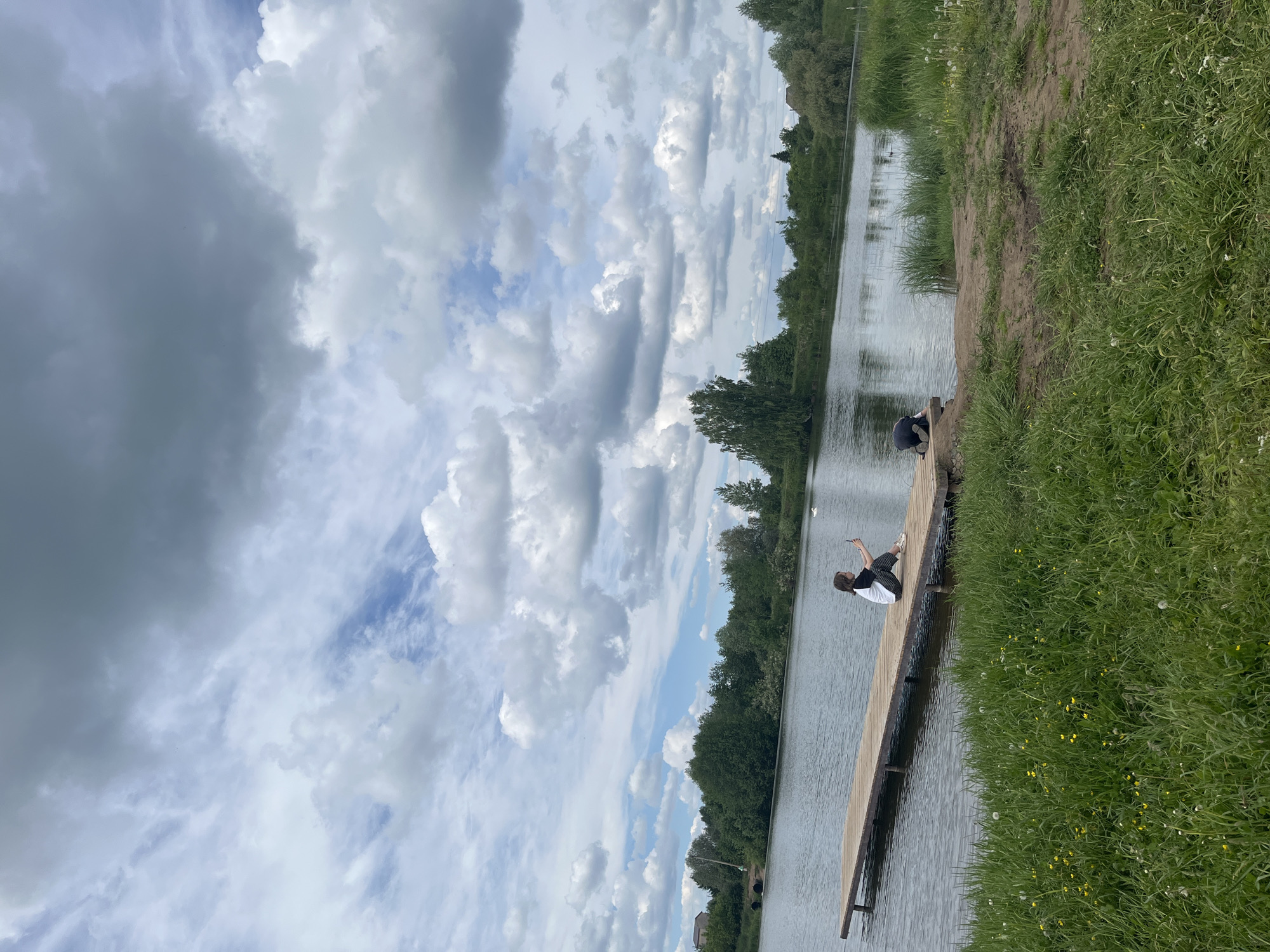 The image captures a peaceful outdoor scene by a body of water in Фоки, Russia, likely a lake or a wide river, under a cloudy sky. The photograph is oriented vertically, but depicts a landscape view when corrected.
In the foreground, a wooden pier extends from a grassy bank into the water. Two individuals are visible on the pier. The person closer to the camera is positioned on the left side of the pier, wearing a dark top and lighter bottoms, and appears to be crouching or leaning over the edge towards the water. Further down the pier, a second person is also crouching or sitting, wearing a white short-sleeved top and dark striped pants. This individual holds a small white object, possibly a phone, in their right hand, pointing it towards the water or the other person, suggesting they might be taking a picture or video.
The water reflects the cloudy sky and the distant trees. On the right side of the image, a lush green grassy bank runs along the water's edge, dotted with small yellow flowers. Across the water, the opposite bank is lined with dense green trees and foliage, forming a natural horizon.
The sky is predominantly covered with a mix of large, fluffy white cumulus clouds and some darker, grey clouds, indicating overcast but not stormy weather. Patches of clear blue sky are visible between the clouds, suggesting it is daytime. No text is visible in the image. The overall impression is one of calm and relaxation in a natural environment.