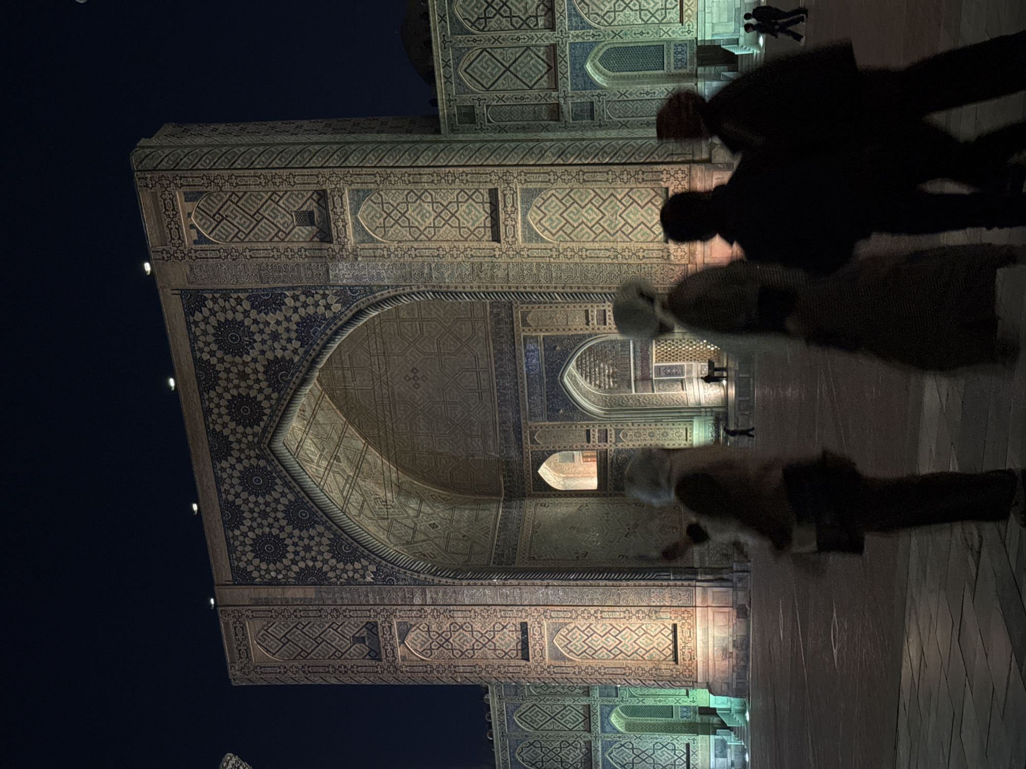 The image depicts the entrance to a building with intricate tilework, likely a religious or historical structure. The location is likely in Samarkand City, Uzbekistan, given the architectural style. The building is illuminated with warm lighting, contrasting with the dark sky. There are people walking around the building, some appearing blurred due to movement, suggesting the photo was taken at night with a slow shutter speed. The architectural details are a prominent feature, showcasing complex geometric patterns and designs.