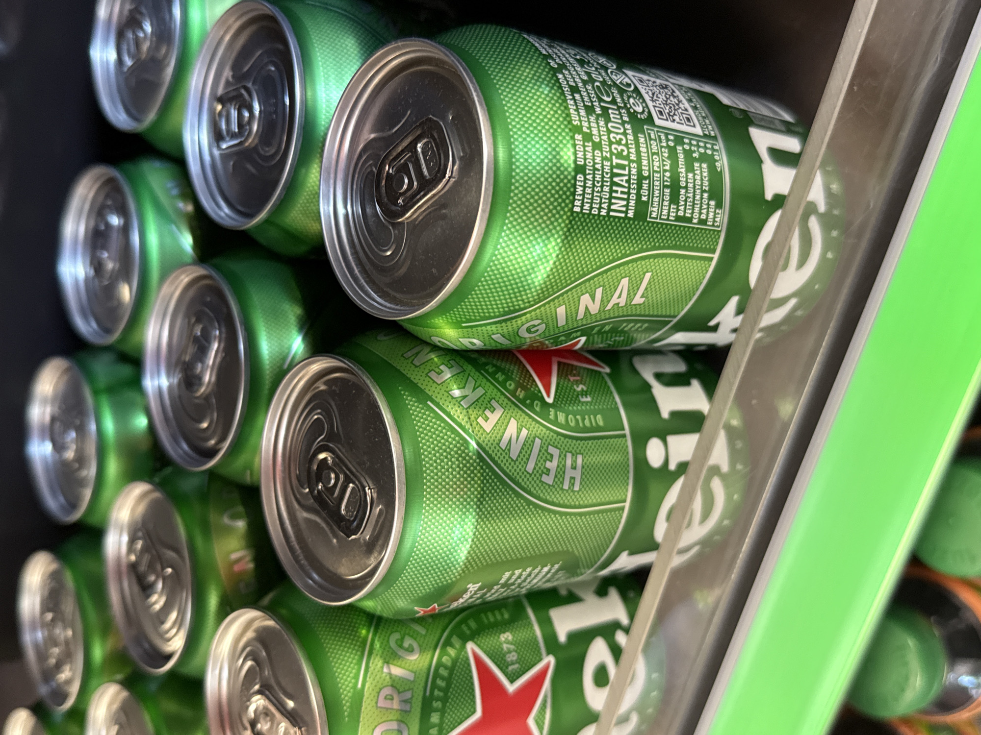 A close-up, eye-level shot shows a display of green Heineken beer cans in a cooler. The cans are packed tightly together, with their silver tops visible. The lighting is somewhat dim, highlighting the shiny surfaces of the cans. In the foreground, a bright green shelf is visible, adding a splash of color. The background is dark and blurred, putting the focus entirely on the beer cans. The scene is consistent with a retail environment, such as a supermarket or convenience store. There are no people or specific identifiable landmarks that would indicate the location within Bad Lauterberg im Harz, Germany, beyond it being a commercial setting likely within the city.