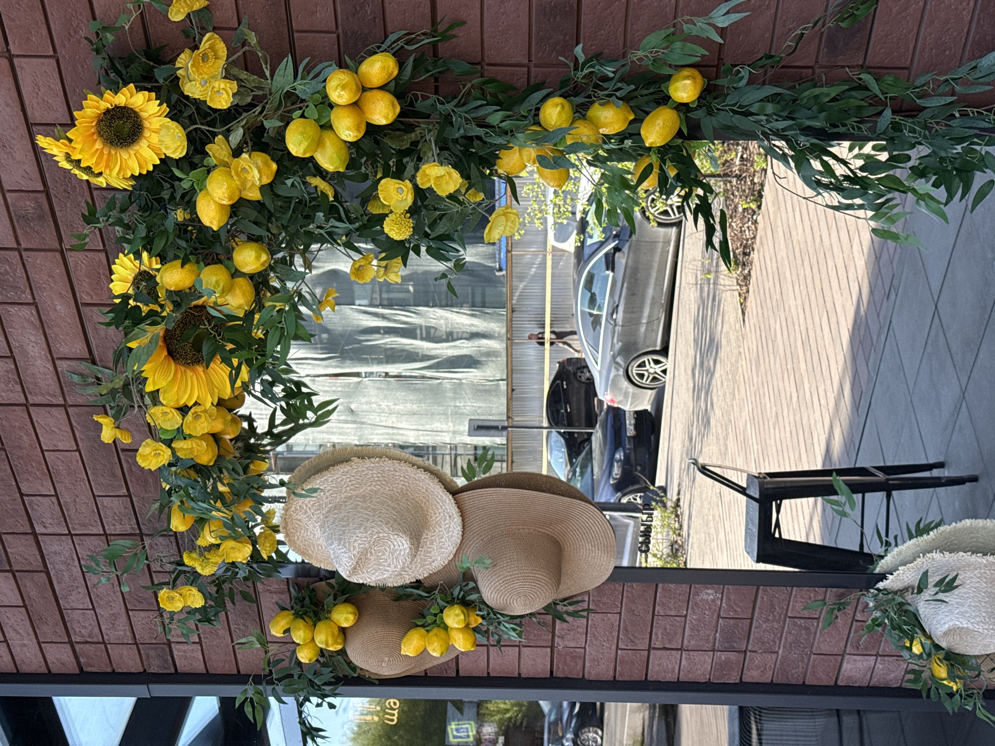 The image captures a decorative display on a brick building in Krasnoyarsk, Russia. The building has a dark frame outlining a window or storefront. Adorning the facade is an arrangement of artificial sunflowers, yellow flower clusters, faux lemons, and green foliage, forming a semi-arch shape. Three straw hats are incorporated into the display. The scene reflects a street view including parked cars and a pedestrian walkway with a metal chair. Reflections in the window or glass storefront reveal the outdoor environment.