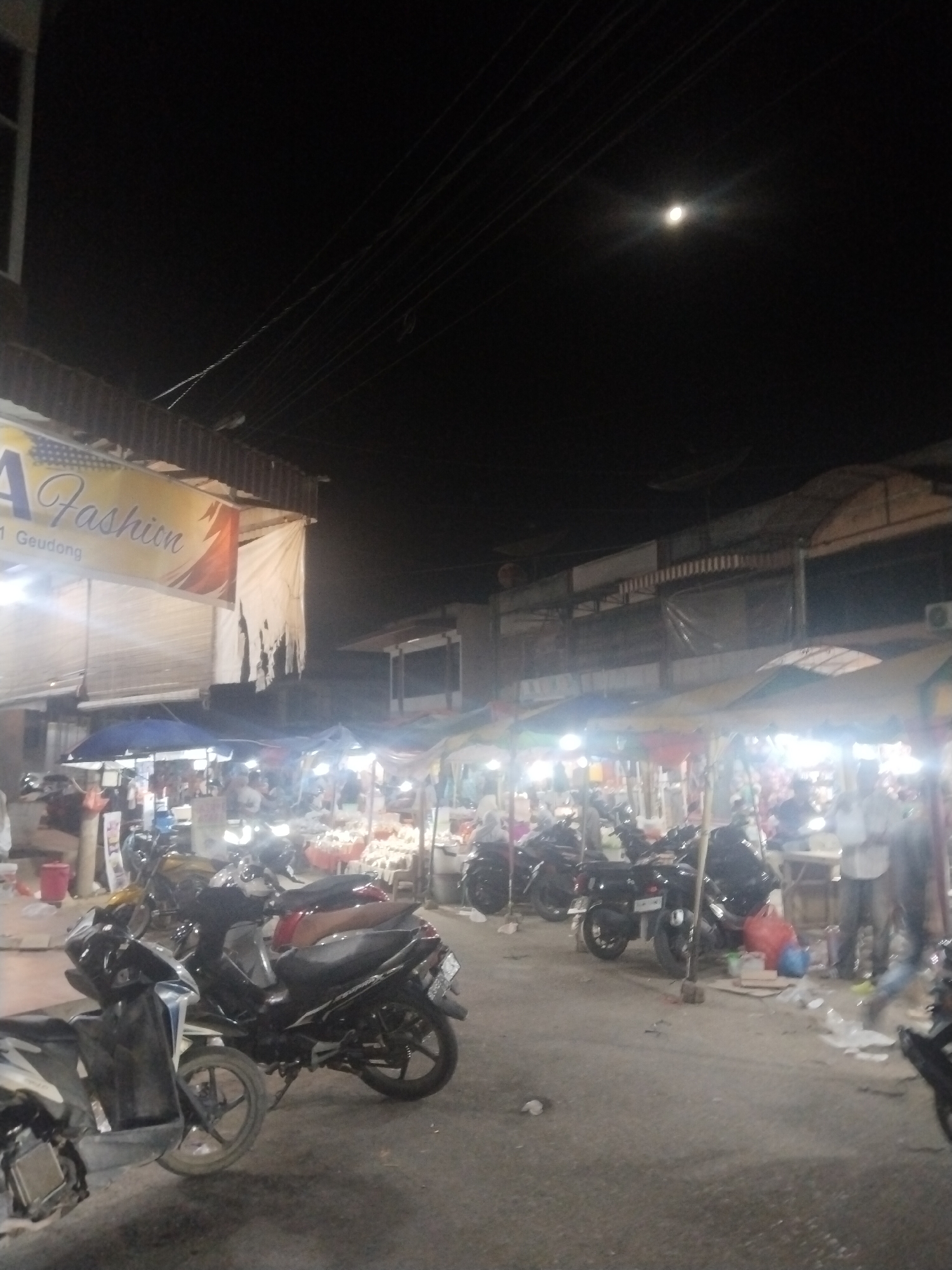 The image depicts a street scene at night in Keudegeudong, Indonesia. The street is lined with market stalls covered by tarpaulins, illuminated by bright lights. A banner for "A Fashion" is visible on a building to the left. Several parked motorcycles are in the foreground. The sky is dark, with a bright moon visible. People can be seen standing and milling around the market stalls. Trash is scattered on the ground. Power lines are strung across the sky.