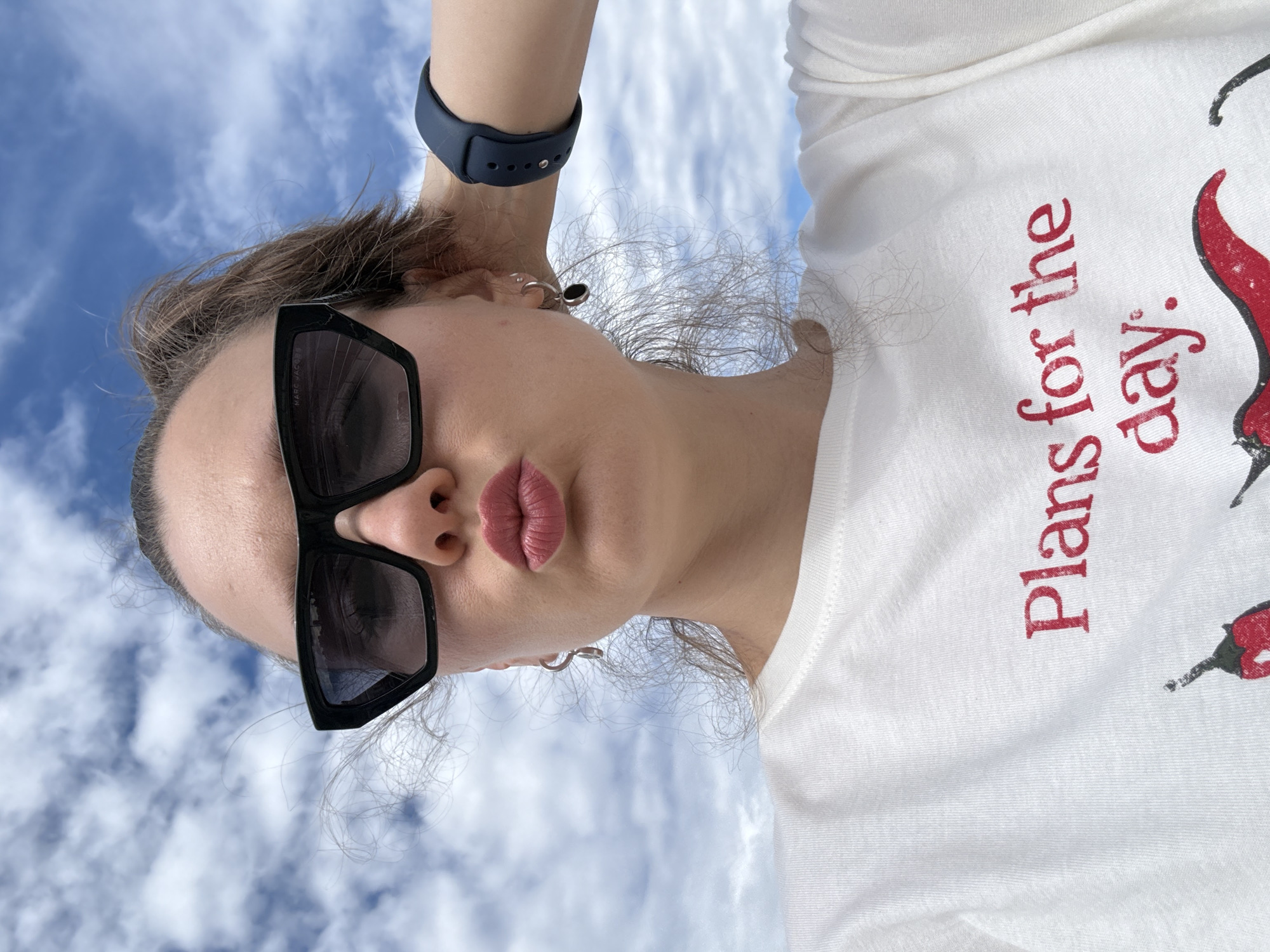 This is a low-angle selfie of a young woman with her arm raised, her hand behind her head. She is wearing large, black, rectangular sunglasses and a white t-shirt with red text that reads "Plans for the day." She has her lips pursed. A dark blue fitness tracker is visible on her wrist. Her hair is light brown and appears windblown. She is wearing a small hoop earring. The background is a bright blue sky with scattered white clouds, suggesting it is daytime and the weather is pleasant. The scene appears to be outdoors, possibly in a park or open area. The image is oriented with the sky at the top, implying the photograph was taken looking upwards. The style of the t-shirt design and the general aesthetic suggest a casual, relaxed mood.