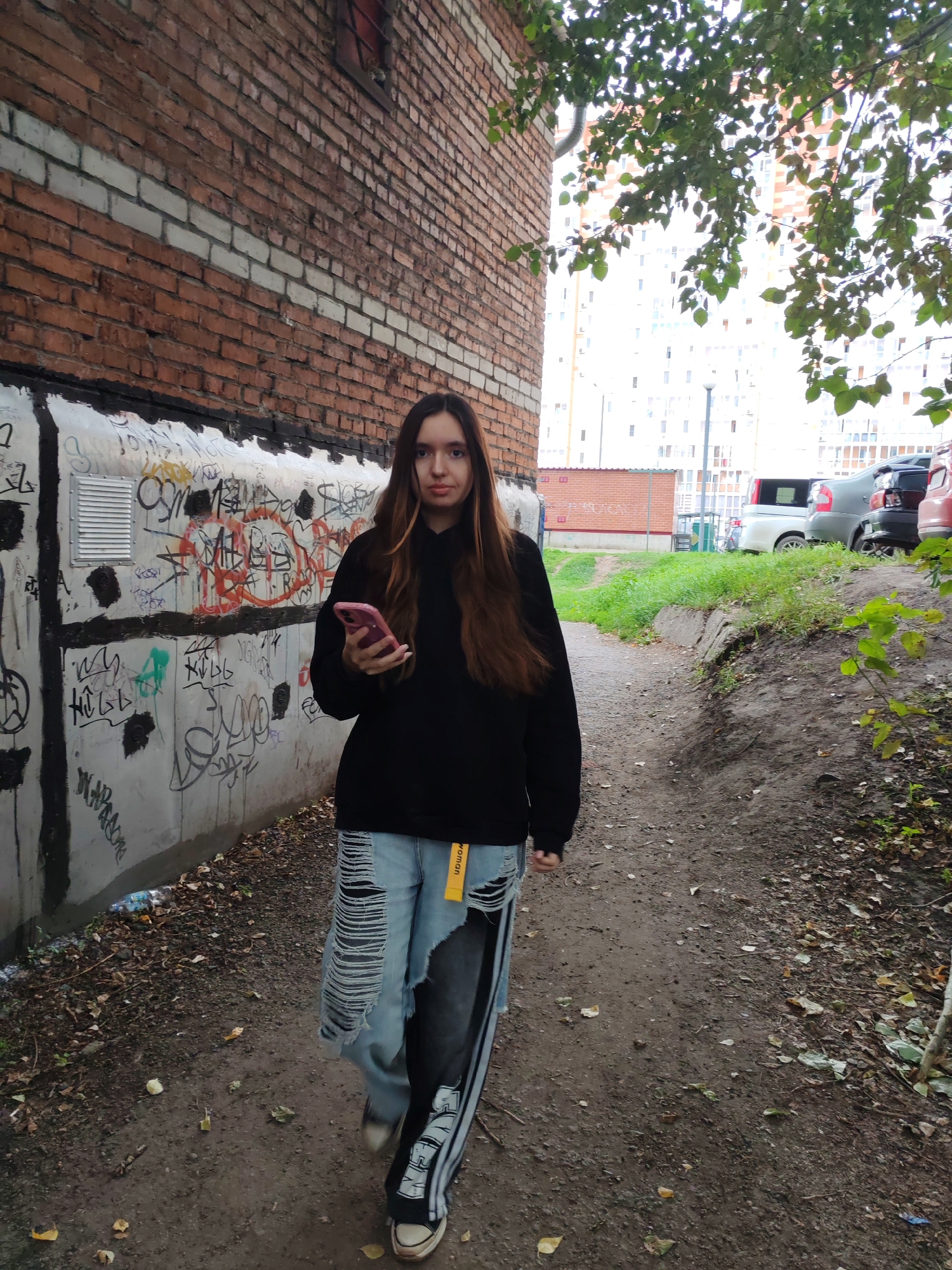 A young woman walks down a dirt path in front of a graffiti-covered wall and a brick building. She is wearing a black hoodie, ripped denim jeans, and sneakers. She is holding a pink smartphone in her right hand. The path is lined with trees and grass. In the background, there are parked cars and apartment buildings.