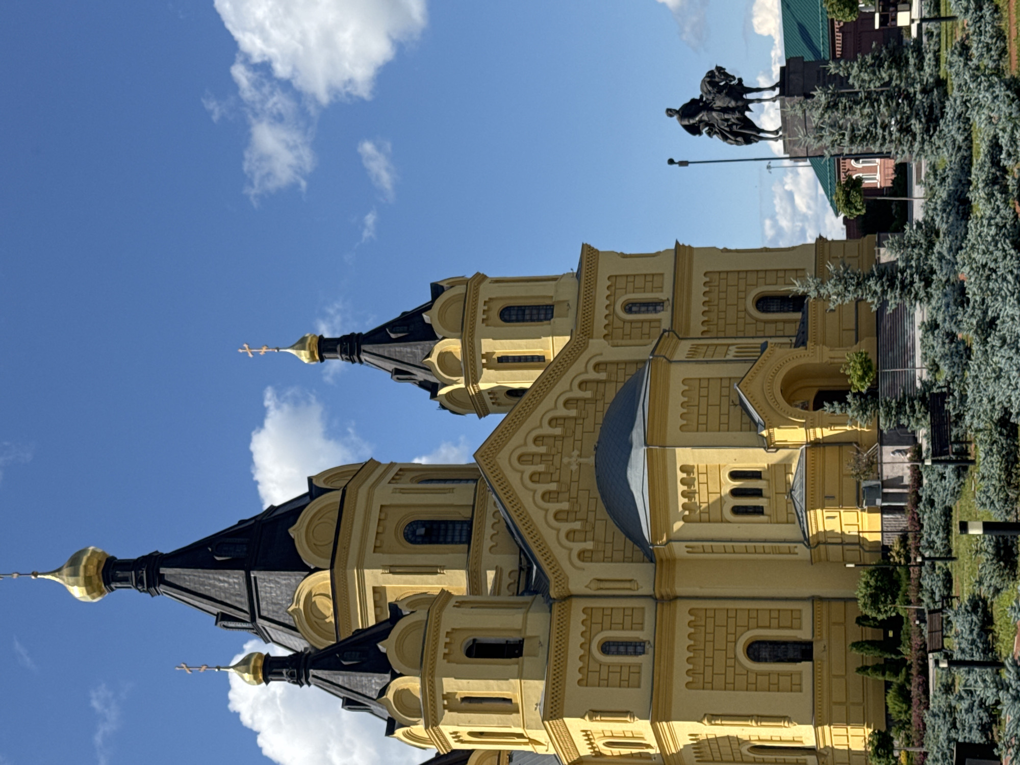 The image depicts a grand yellow cathedral with multiple domes and spires, a common architectural style for Orthodox churches, set against a vibrant blue sky with scattered white clouds. In front of the cathedral, a dark bronze equestrian statue of a man on horseback stands on a pedestal. The surrounding area is a well-maintained park with manicured bushes and trees, interspersed with benches and lampposts. The cathedral's facade is intricately decorated with architectural details such as arched windows and decorative cornices. The overall scene suggests a public space, likely a historical or religious site in Nizhny Novgorod, Russia. The cathedral's prominence and the statue's presence indicate a location of significant cultural or historical importance within the city.