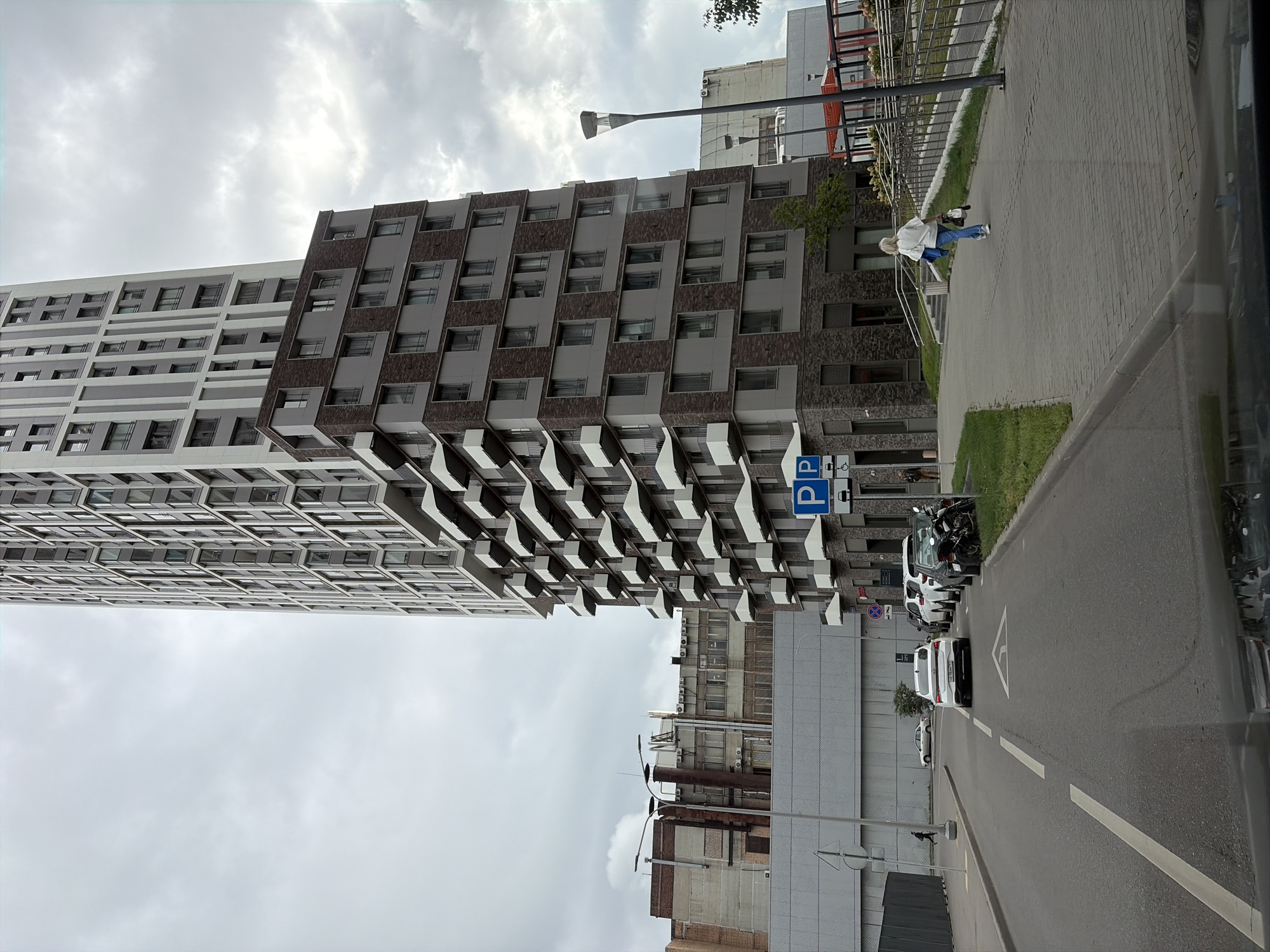 The image shows a cloudy day in Moscow, Russia, with a large, modern apartment building as the focal point. The building is tall and features a distinctive design with numerous protruding balconies or terraces. To the left of the apartment building, there is an older, industrial-style building with brick and concrete elements. A street runs in front of these buildings, with several cars parked along the side and one driving away. A pedestrian walkway with a railing is visible to the right of the street, with a woman walking away from the camera. Parking signs are posted near the walkway. The overall scene depicts an urban environment with a mix of contemporary and older architecture.