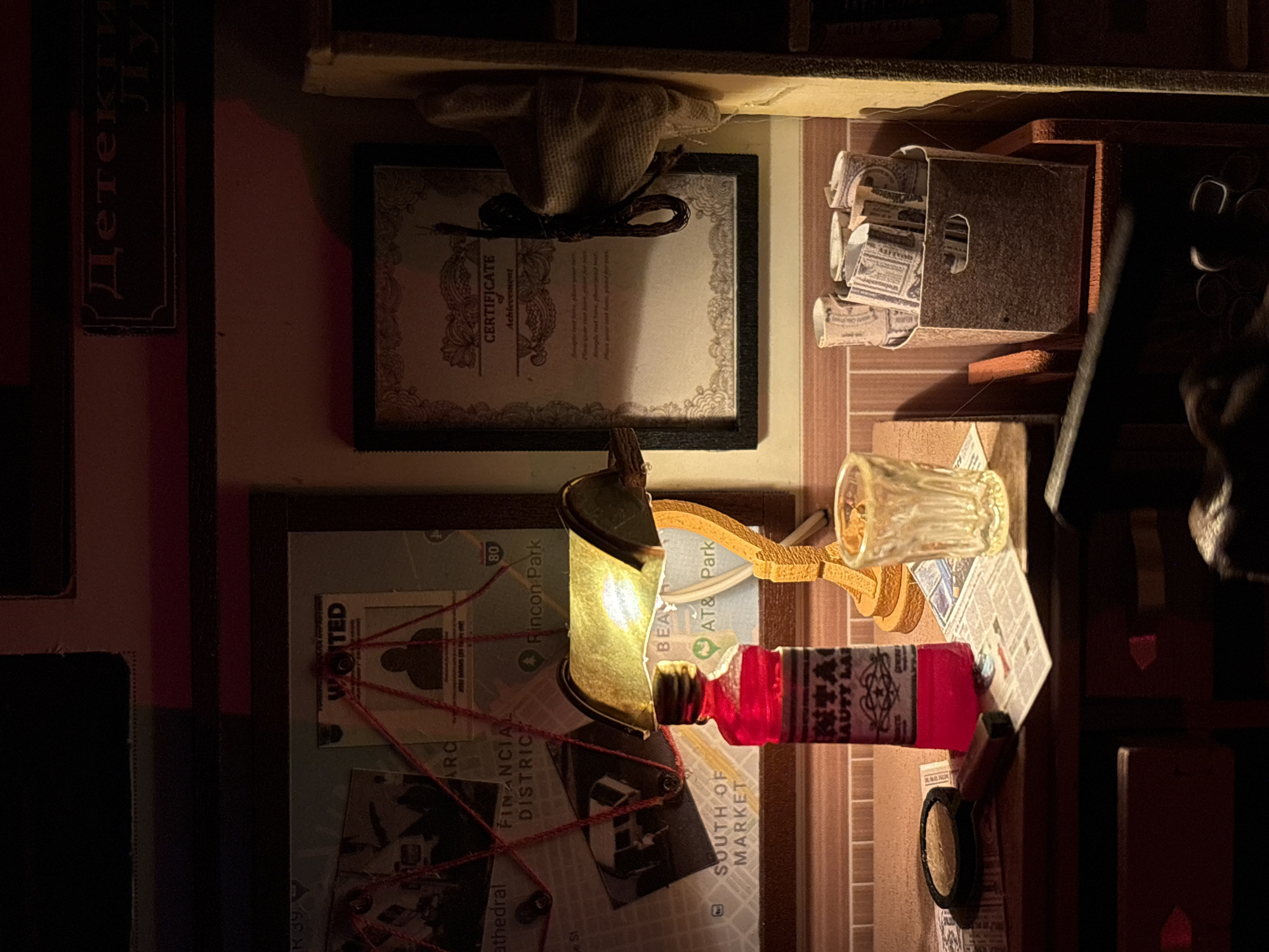 The image depicts a miniature detective's desk in an indoor setting, likely an office. The desk is cluttered with various items suggestive of an investigation. A map of San Francisco, annotated with red string connecting points of interest like "Financial District" and "Rincon Park," is pinned to a corkboard. A "WANTED" poster with a silhouetted figure is also displayed.
On the desk, a red bottle, possibly containing liquid, stands beside a glass of water. A magnifying glass rests on a newspaper. Rolled-up bills, bundled together in a small basket, suggest a monetary aspect to the case. A framed certificate, with the word "CERTIFICATE" prominently displayed, hangs on the wall. The lighting is dim, with a desk lamp casting a warm glow on the scene, creating a noir atmosphere. The overall impression is that of a meticulous, perhaps even obsessive, detective at work.
