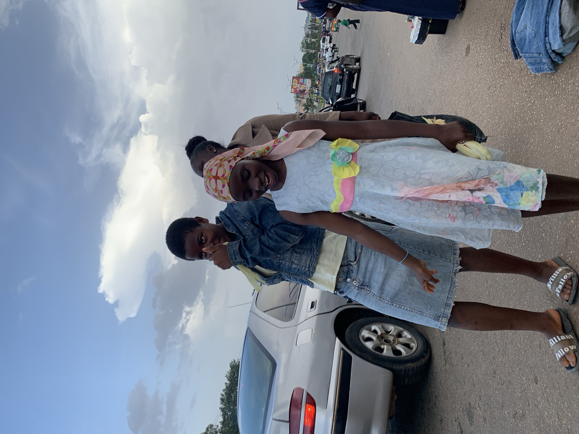 The image shows two young girls on a street in Jos, Nigeria. The girl on the left is wearing a denim jacket, a t-shirt, and a denim skirt. She is giving a thumbs-up gesture. The girl on the right is wearing a light blue dress with a floral pattern and a pink headscarf. She is smiling. Behind them, a silver car is parked on the side of the road. In the background, there is traffic, streetlights, and buildings. The sky is cloudy.