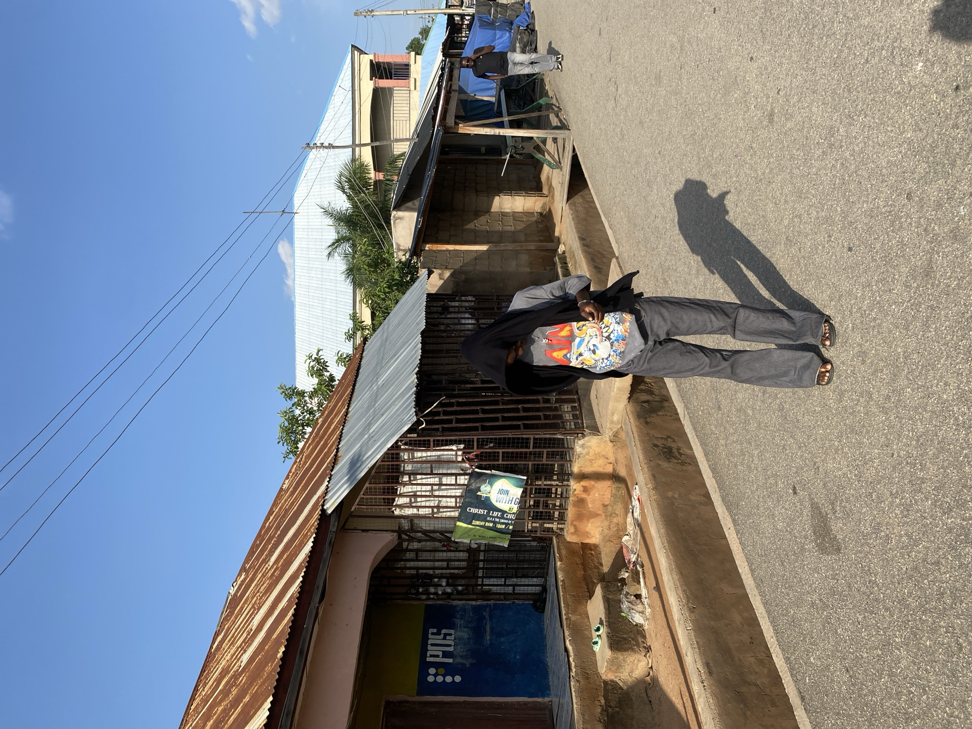 The image shows a street scene in Jalingo, Nigeria. In the foreground, a person wearing a black hijab and a grey t-shirt with a cartoon graphic is walking away from the camera, facing slightly to their right. They are also wearing loose grey pants and sandals. To the left of the person, there is a building with a corrugated iron roof and a blue facade with "POS" written on it in white lettering. A metal gate is in front of the building. To the right of the foreground person, further down the street, another person in a black shirt and grey pants is walking towards the camera, talking on a mobile phone. The street is paved and appears to be a commercial area with various shops and structures lining both sides. The sky is clear blue with a few scattered clouds. The overall environment suggests a warm, sunny day in a bustling urban setting.