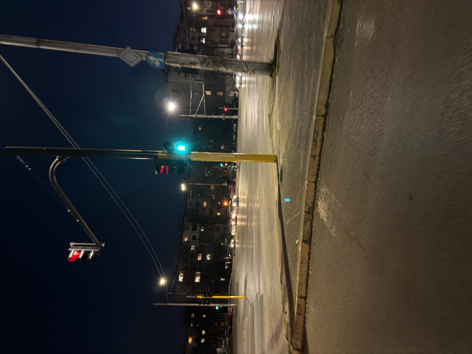 This night-time image depicts an urban intersection within Sofia, Bulgaria, illuminated by artificial light sources against a dark blue sky. In the immediate foreground, a paved road surface transitions to a concrete curb and an adjacent paved area. A small blue plastic item and white debris are visible on the ground near the curb.
A prominent yellow pole stands in the center-right of the foreground, supporting a vertical traffic light assembly. The lower signal displays a green light, while an upper segment displays a red light, likely for a separate turning lane or direction. An overhead traffic light is visible above the intersection, displaying a red signal. To the right of the yellow pole, a taller, grey utility pole supports two road signs: a diamond-shaped priority road sign (white background with a blue border) and, beneath it, a blue square sign with a white capital 'A' and a diagonal white arrow pointing down-right. Below this, a white square sign with a black numeral '4' is affixed.
The multi-lane intersection beyond is occupied by multiple vehicles, whose headlights and taillights are illuminated, appearing to be stationary or moving slowly in various lanes. In the background, several multi-story apartment buildings with numerous illuminated windows are visible, indicative of a residential area. No individuals are present in the visible scene.