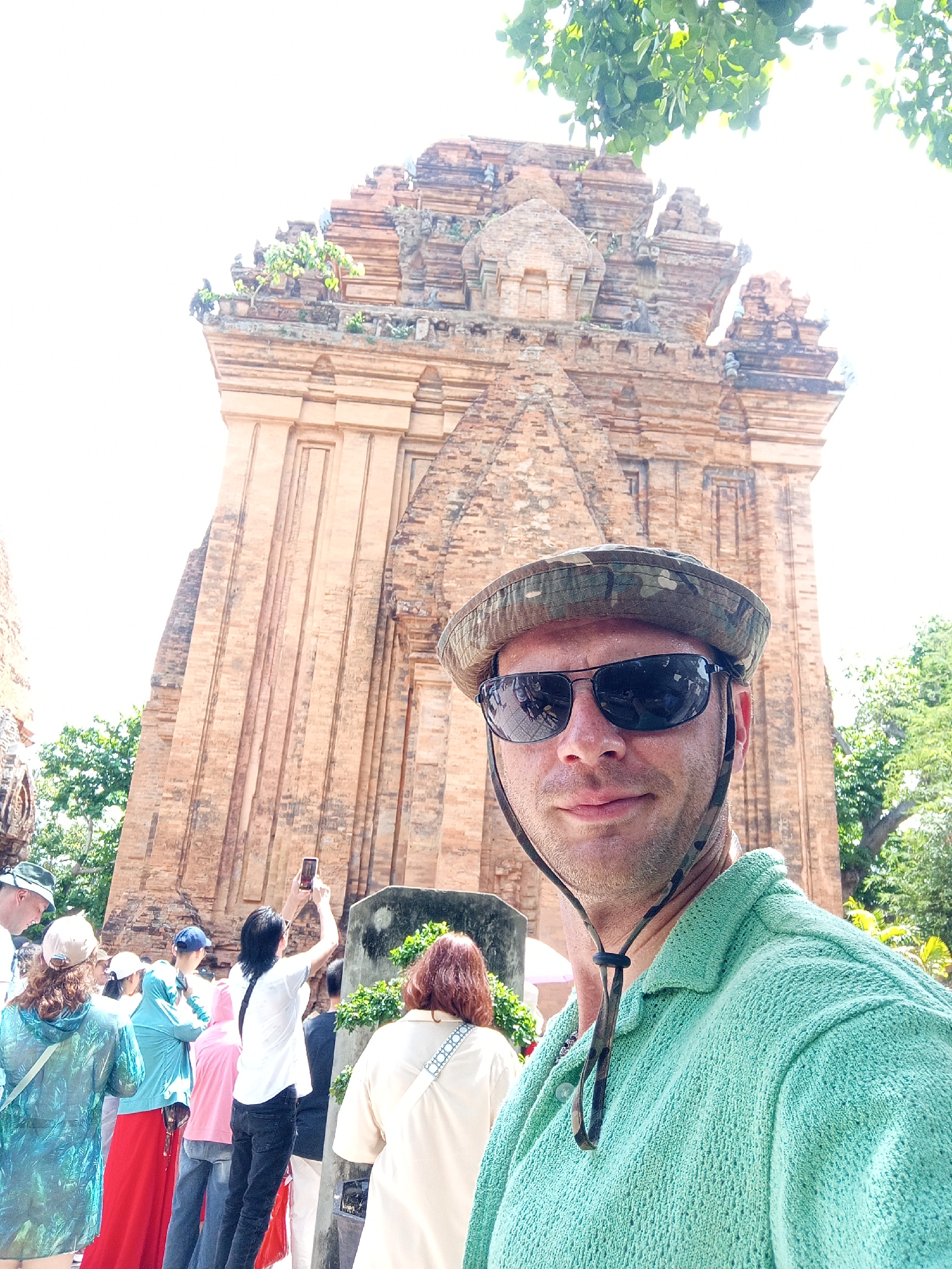 A man wearing a camouflage hat and sunglasses takes a selfie in front of a brick tower, possibly a Po Nagar Cham Tower, in Nha Trang, Vietnam. He is wearing a light green, textured polo shirt. Behind him, a group of people are visible, some holding up phones to take pictures. The tower is made of red brick and has several tiers and decorative elements. Lush green trees are visible in the background, and the sky is bright and washed out, indicating a sunny day. The overall scene suggests a tourist visiting a historical site.