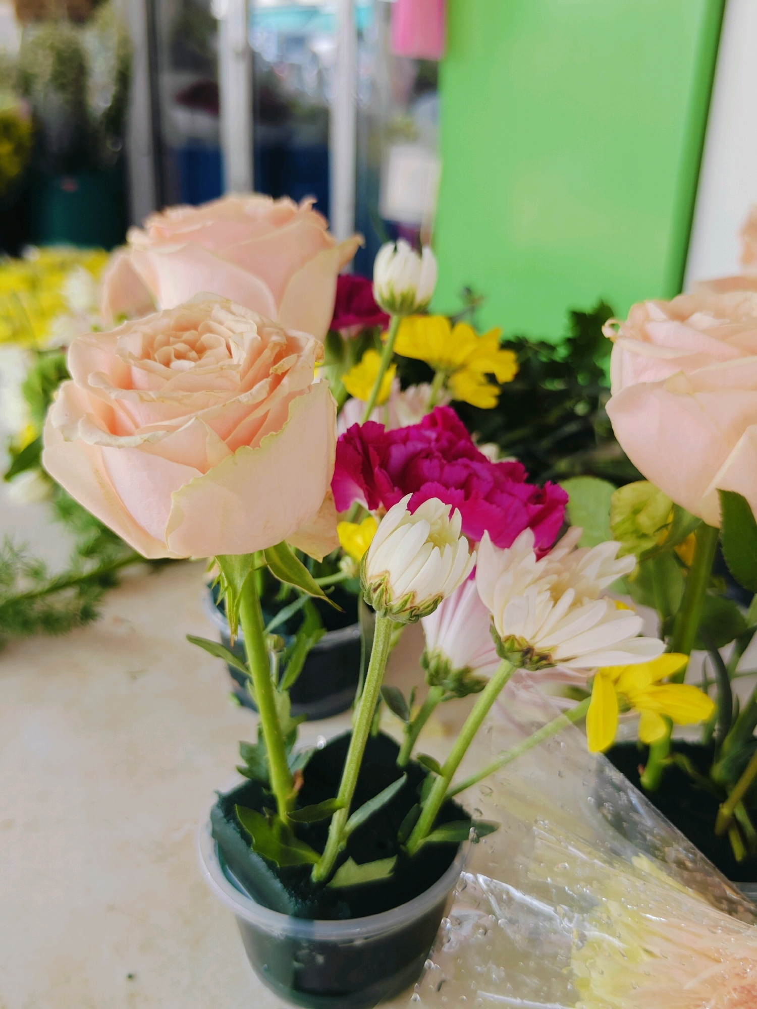 This is a close-up, eye-level, indoor photograph of a floral arrangement, likely taken at a flower shop in Bangkok, Thailand, given the context provided. The arrangement features several light pink roses as the focal point, with their petals unfurling delicately. Interspersed are smaller white daisy-like flowers and bright yellow flowers, adding pops of color and texture. A few deep pink or magenta carnations are also visible, providing a darker contrast. The flowers are arranged in small, clear plastic cups filled with dark green floral foam. The background is softly blurred, showing hints of other floral displays and a bright green surface, possibly a counter or a wall. The overall lighting is soft and diffused, creating a gentle and appealing aesthetic. The image appears to be taken from a slightly low angle, looking up at the flowers.