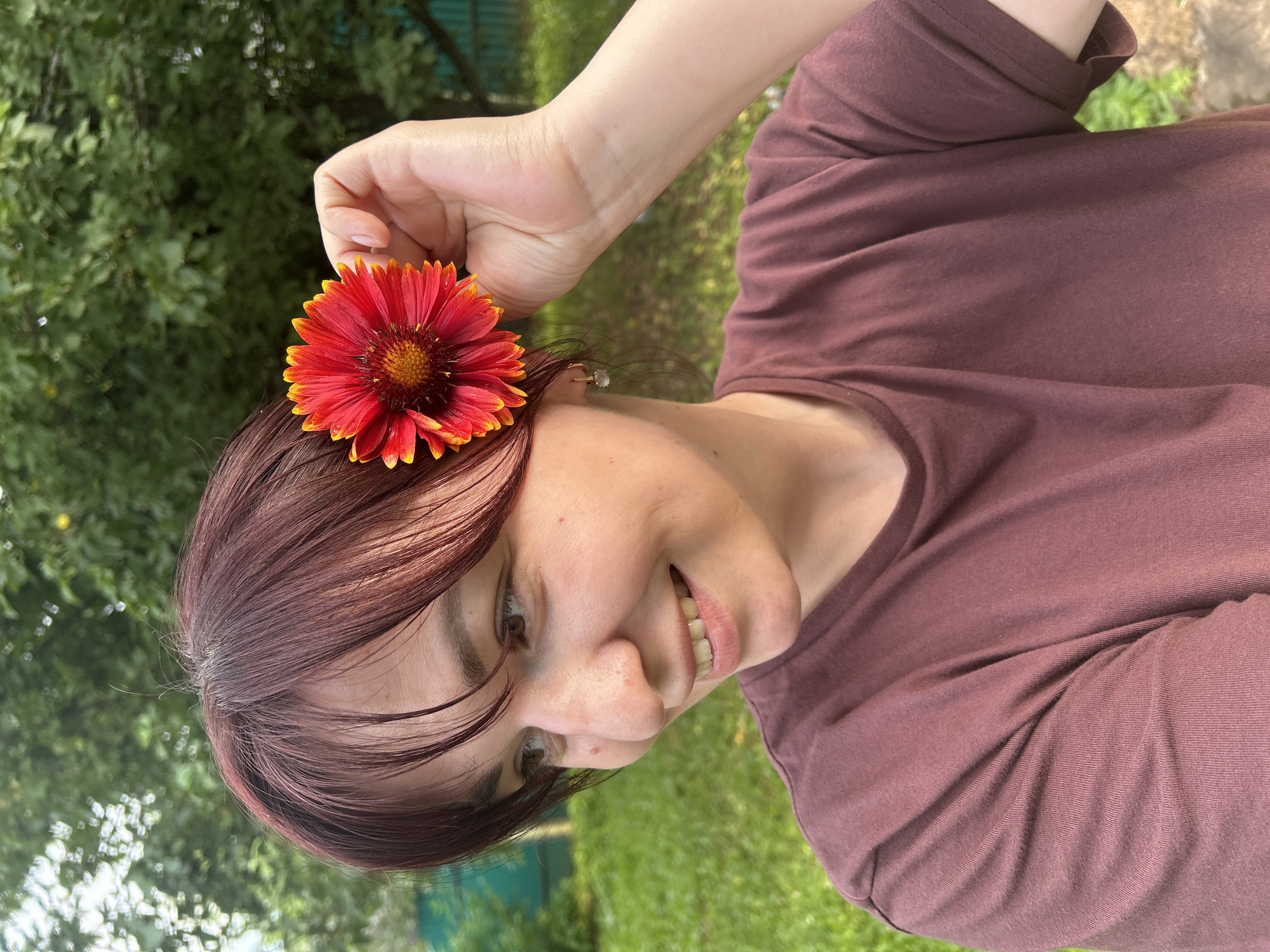The image shows a young person with brown hair, wearing a maroon t-shirt, posing outdoors with a red and yellow flower placed in their hair. The person is smiling and looking towards the camera. The background consists of green foliage and grass, suggesting an outdoor setting, possibly a garden or park. The lighting indicates it is daytime, with natural sunlight illuminating the scene. There are no visible text elements in the image. The overall mood appears cheerful and relaxed. The location context provided is Фоки, Russia.