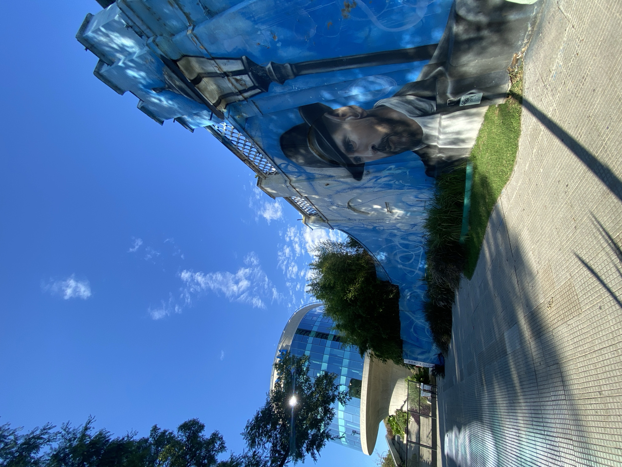 The image shows a mural depicting a man with a hat and a lamppost, painted on the underside of the Pueyrredón Bridge in Buenos Aires, Argentina. The mural has a blue background and is located next to a grassy area and a sidewalk. The sky above the bridge is clear and blue with some white clouds. In the background, a modern glass-fronted building with a curved shape is visible. Trees are also present in the scene. The image appears to be taken during the daytime with bright sunlight, creating shadows on the sidewalk.