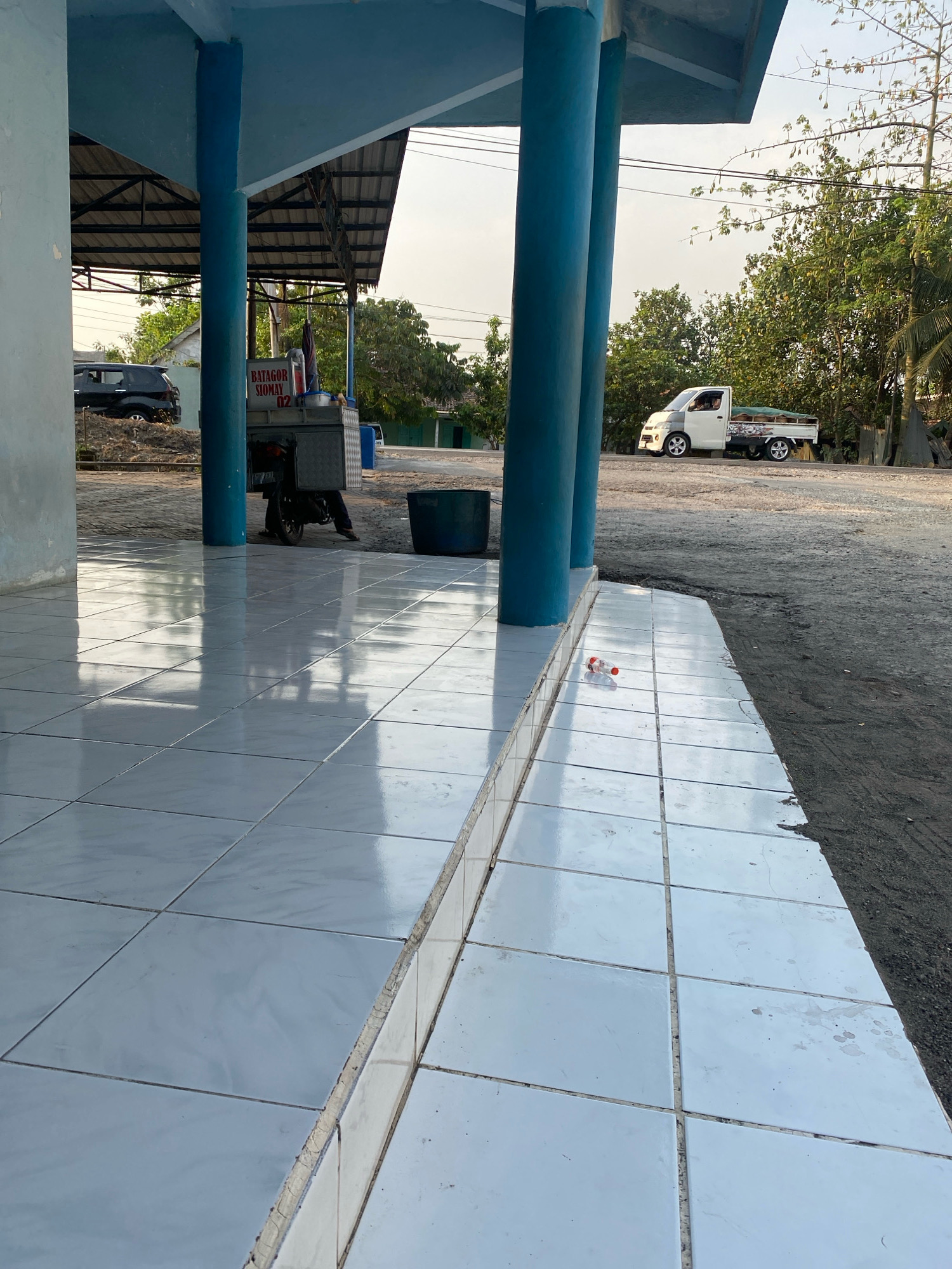 The image depicts a street scene in Kedung Sukodani, Indonesia, in what appears to be late afternoon given the lighting. The foreground is dominated by a tiled walkway, reflecting the overhead structure. This structure is supported by thick blue pillars and a blue roof. To the left, a portion of a light-colored wall is visible.
In the middle ground, a food cart with "BATAGOR SIOMAY 02" signage is parked, with a person seated behind it. The cart is mounted on a motorbike. A black SUV is parked to the left of the food cart, partially obscured by a pillar. Further in the background, a white pickup truck with a load in its bed is driving on a dirt road. Trees and a few structures are visible behind the road. A dark green cylindrical container is placed on the ground near the food cart.
On the tiled walkway in the foreground, a single empty plastic bottle lies on its side. The overall environment suggests an outdoor commercial area or roadside.