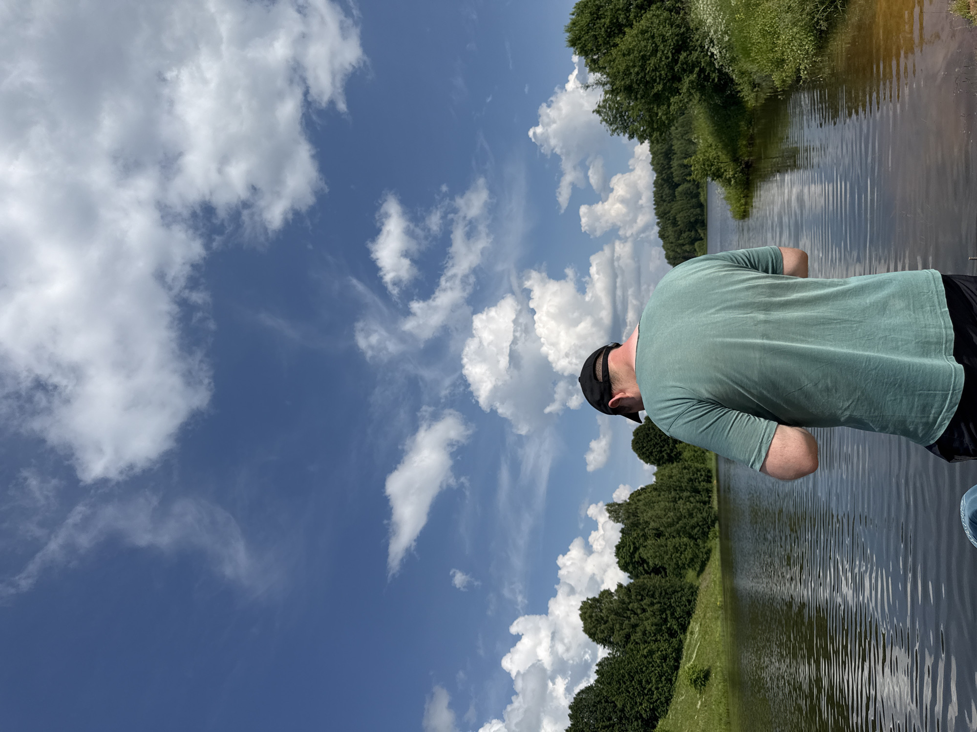 The image depicts a young man wearing a teal t-shirt and a black baseball cap, viewed from behind. He is standing on the edge of a body of water, possibly a lake or river, with his back to the camera. The water is calm with visible ripples, reflecting the bright blue sky dotted with white, fluffy clouds. Lush green trees and vegetation line the opposite bank, forming a natural landscape. The area appears to be a serene, outdoor setting, likely in a rural or park-like environment, consistent with the natural beauty found in and around Zasechnyi, Russia.