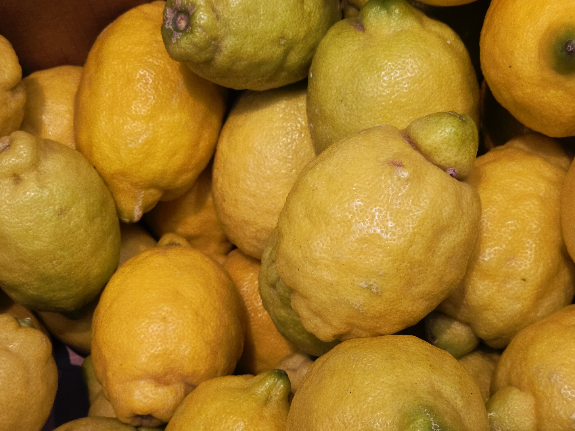 A close-up, high-angle shot shows a collection of lemons. Some are bright yellow, while others are a lighter, greenish-yellow. They are piled together, filling the frame. The location and city are unknown.