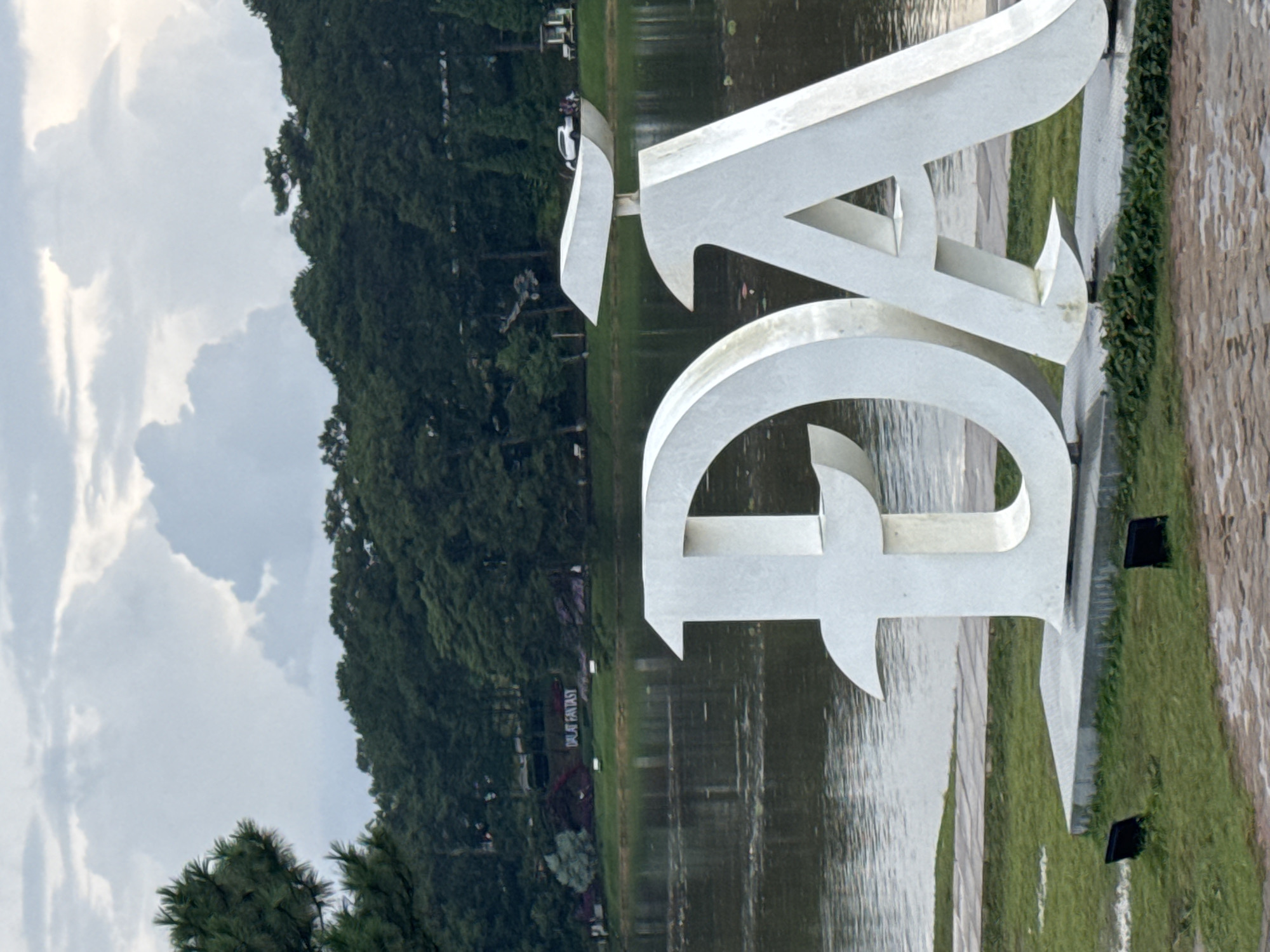 The image shows a large, white, three-dimensional sculpture of Vietnamese letters "Đ" and "A" situated on the edge of a body of water, likely Xuan Huong Lake, in Da Lat, Vietnam. The sculpture is positioned in the foreground on a concrete base surrounded by green grass. In the middle ground, the calm water of the lake reflects the overcast sky. Across the lake, a dense forest of dark green trees covers a hillside, with a sign reading "DALAT FANTASY" visible among the trees in the distance. The sky is cloudy with a mix of light and dark gray formations. The overall scene suggests a serene, natural environment with a prominent landmark.